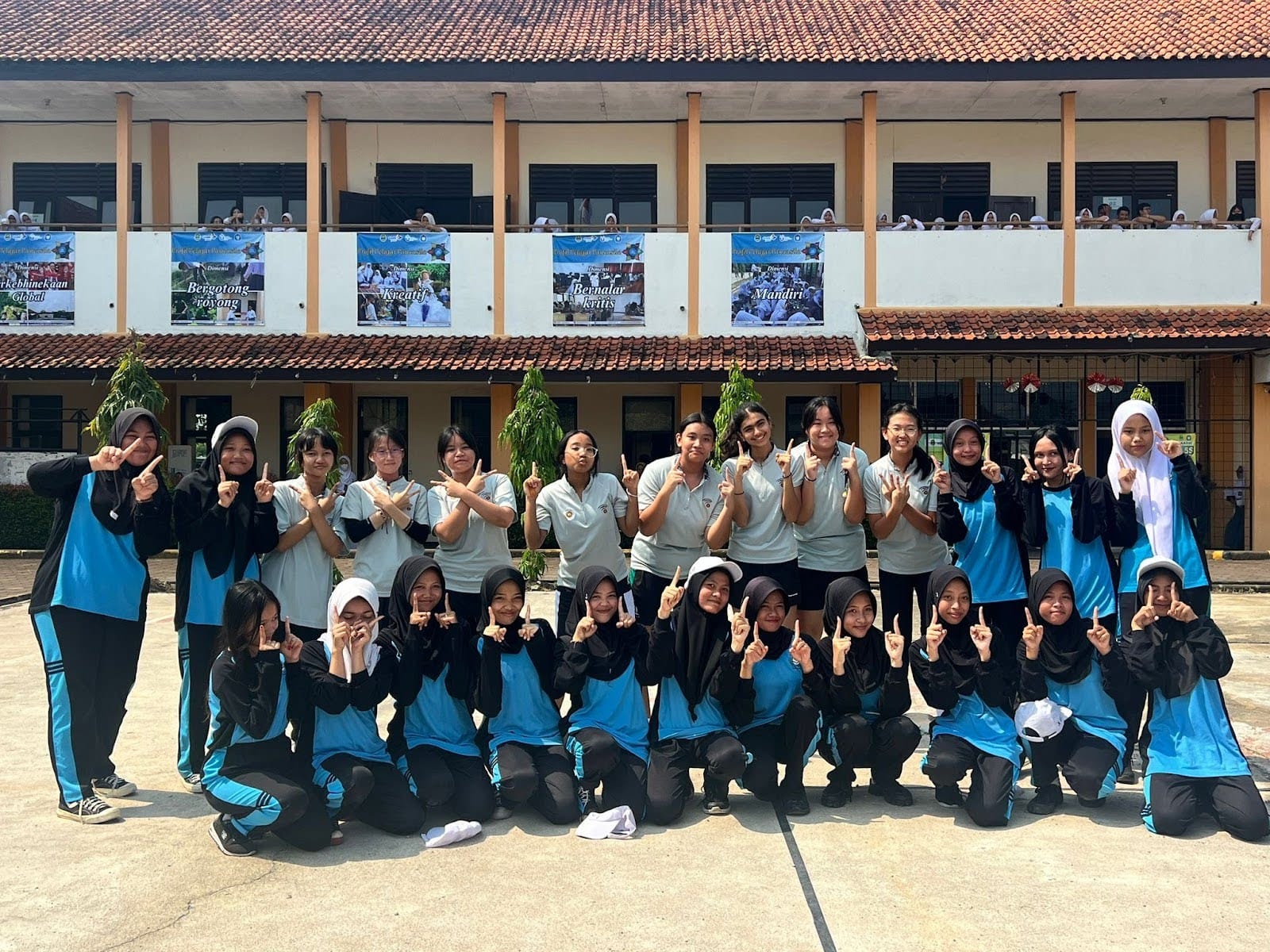 A large group of female students in blue and black uniforms pose with one finger up in a school courtyard.