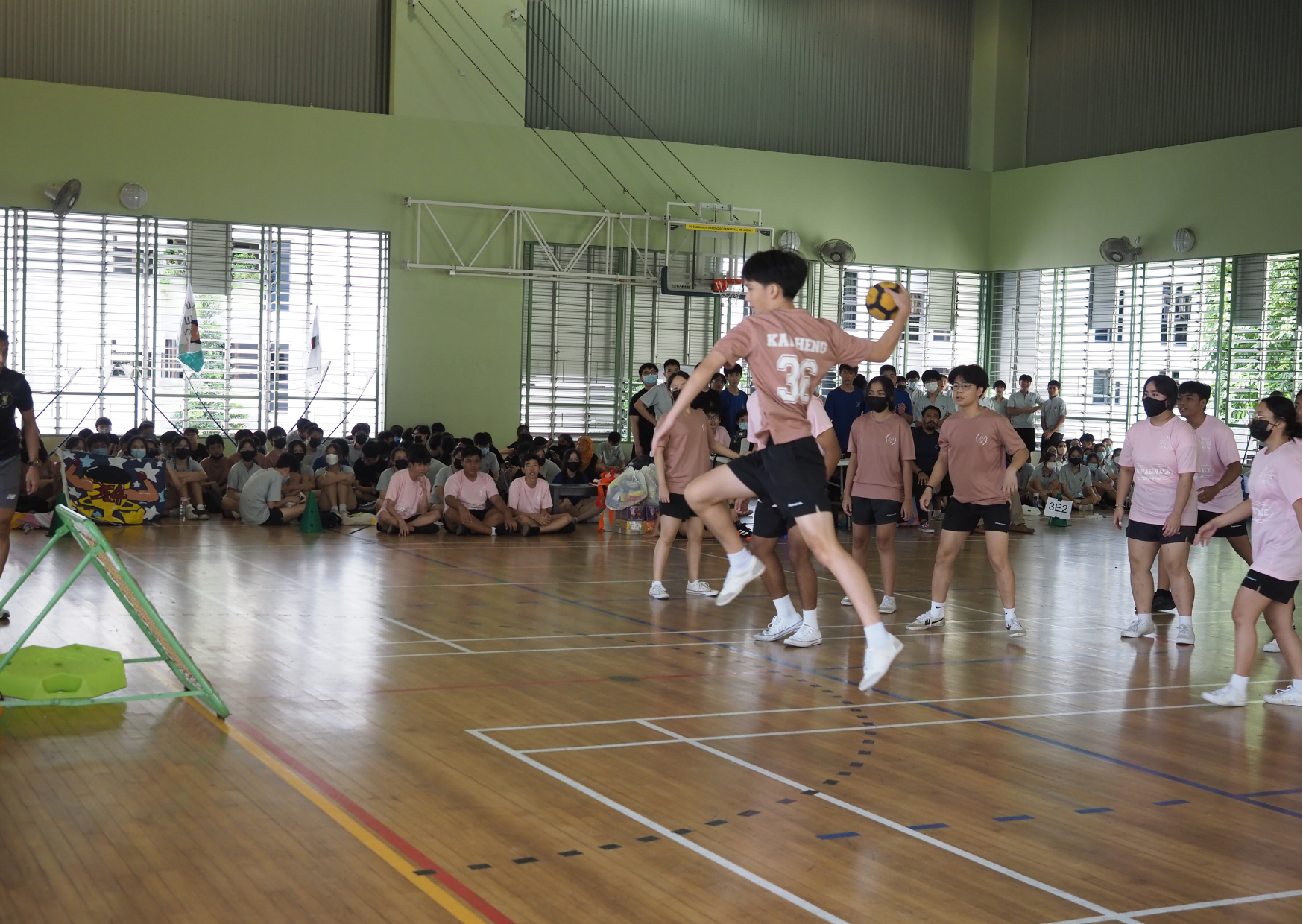 Students play handball in a school gymnasium while many seated students watch from the sidelines.