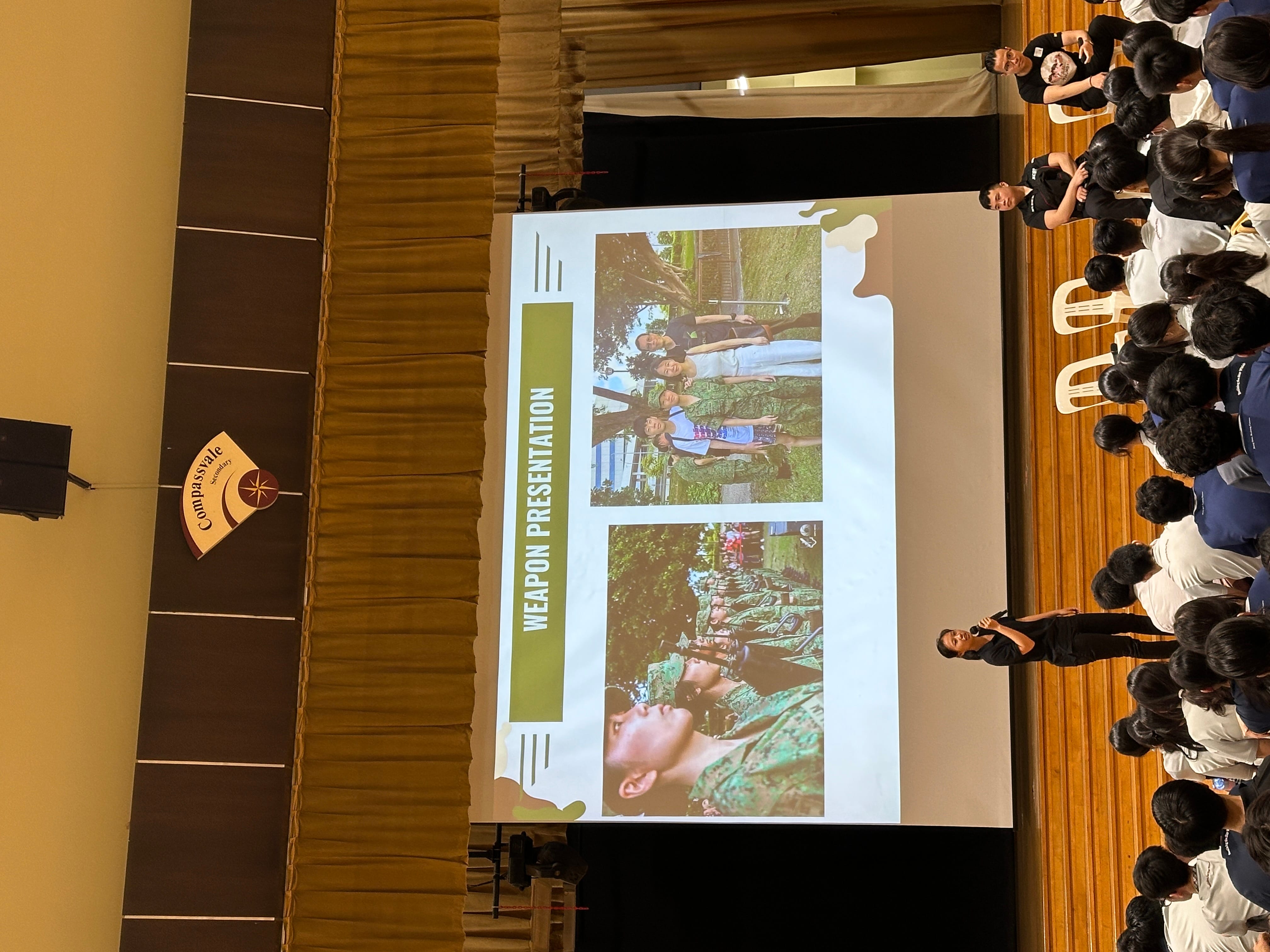 A speaker presents a "Weapon Presentation" slideshow to students in Compassvale Secondary School's hall.