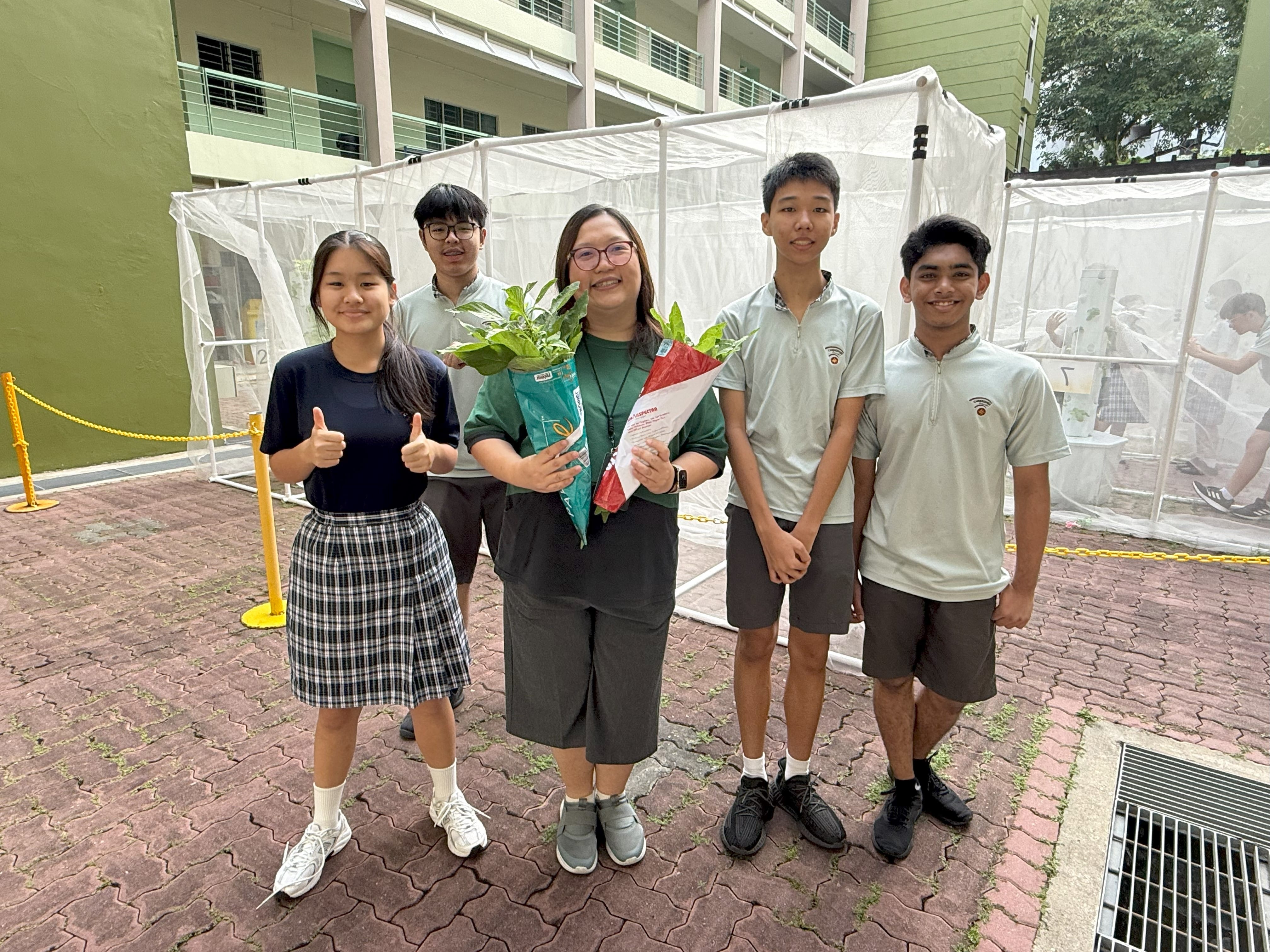 Five people pose outdoors at a school, with one adult holding a potted plant wrapped in red and green paper.