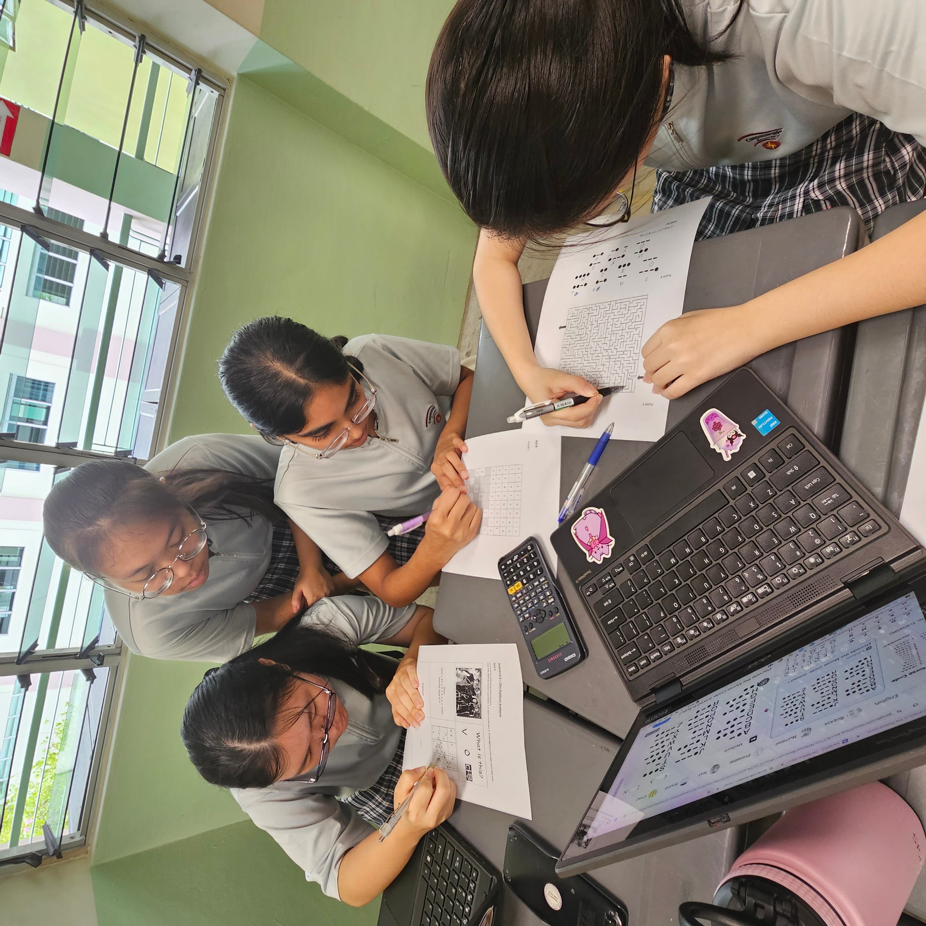 Four uniformed schoolgirls work together at a desk with papers, a laptop, and a calculator near windows.