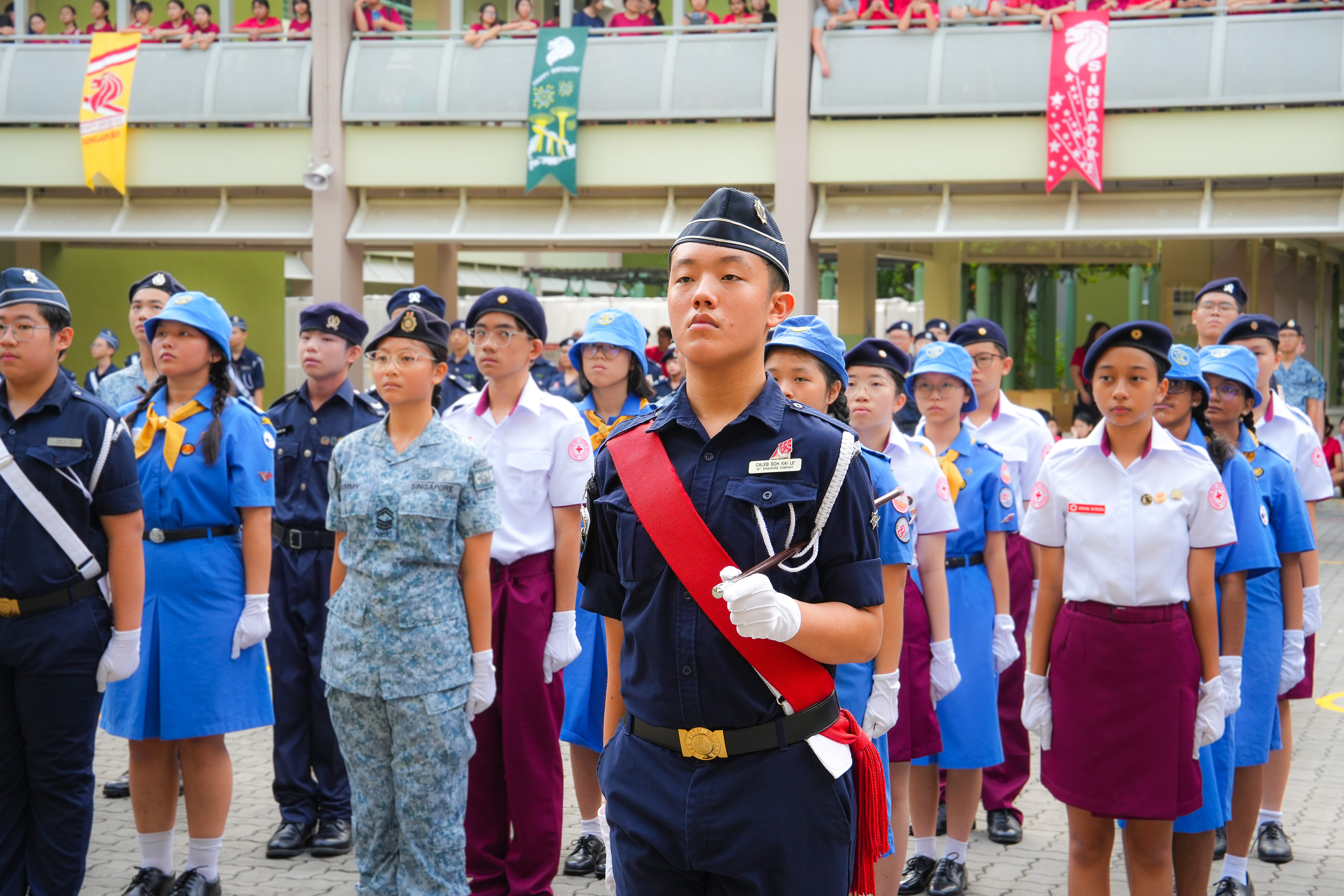 Student in navy uniform with red sash leads a formation of youths in uniforms; crowd watches from balcony