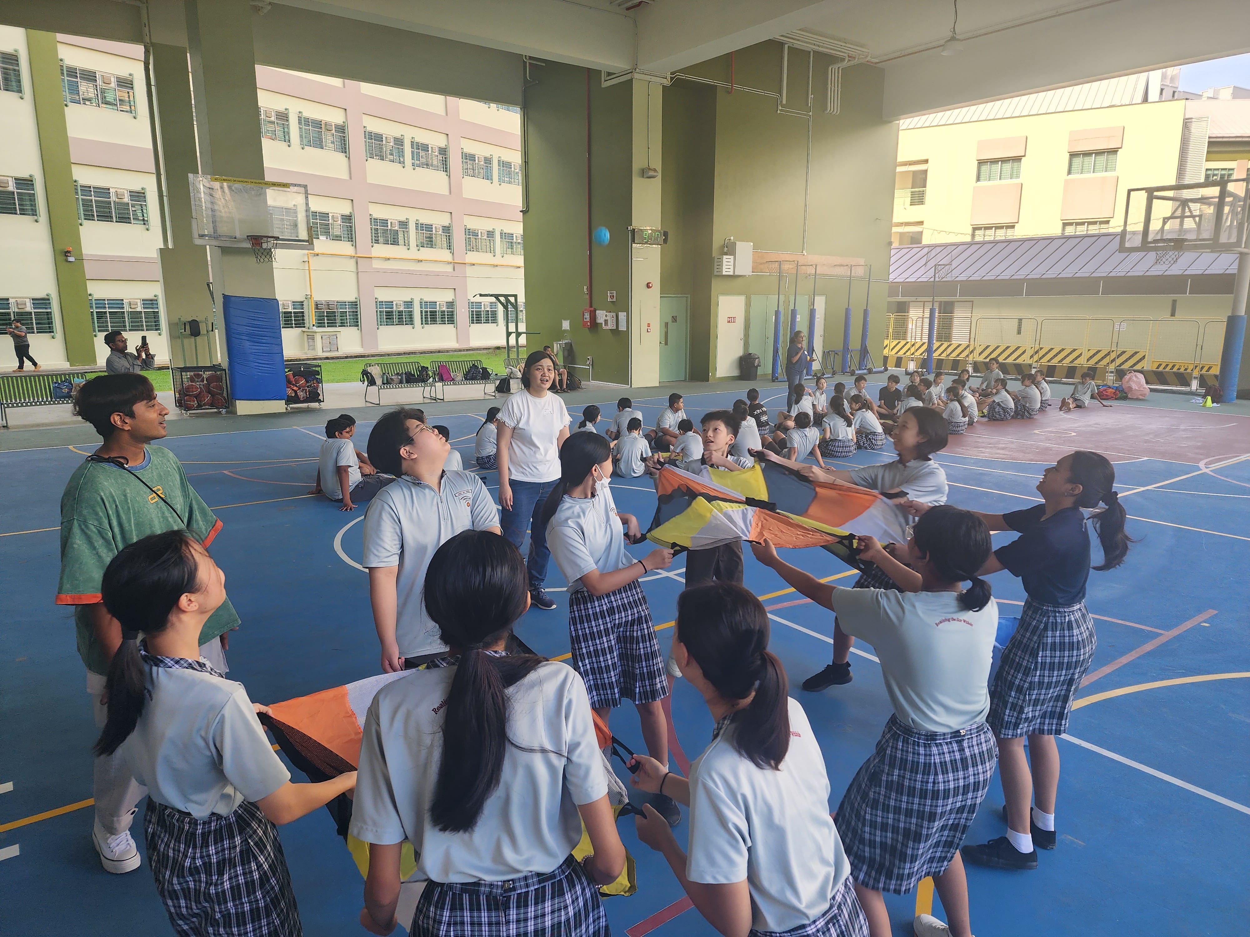 Students in uniforms hold a colorful parachute on an indoor court while others sit in rows behind.