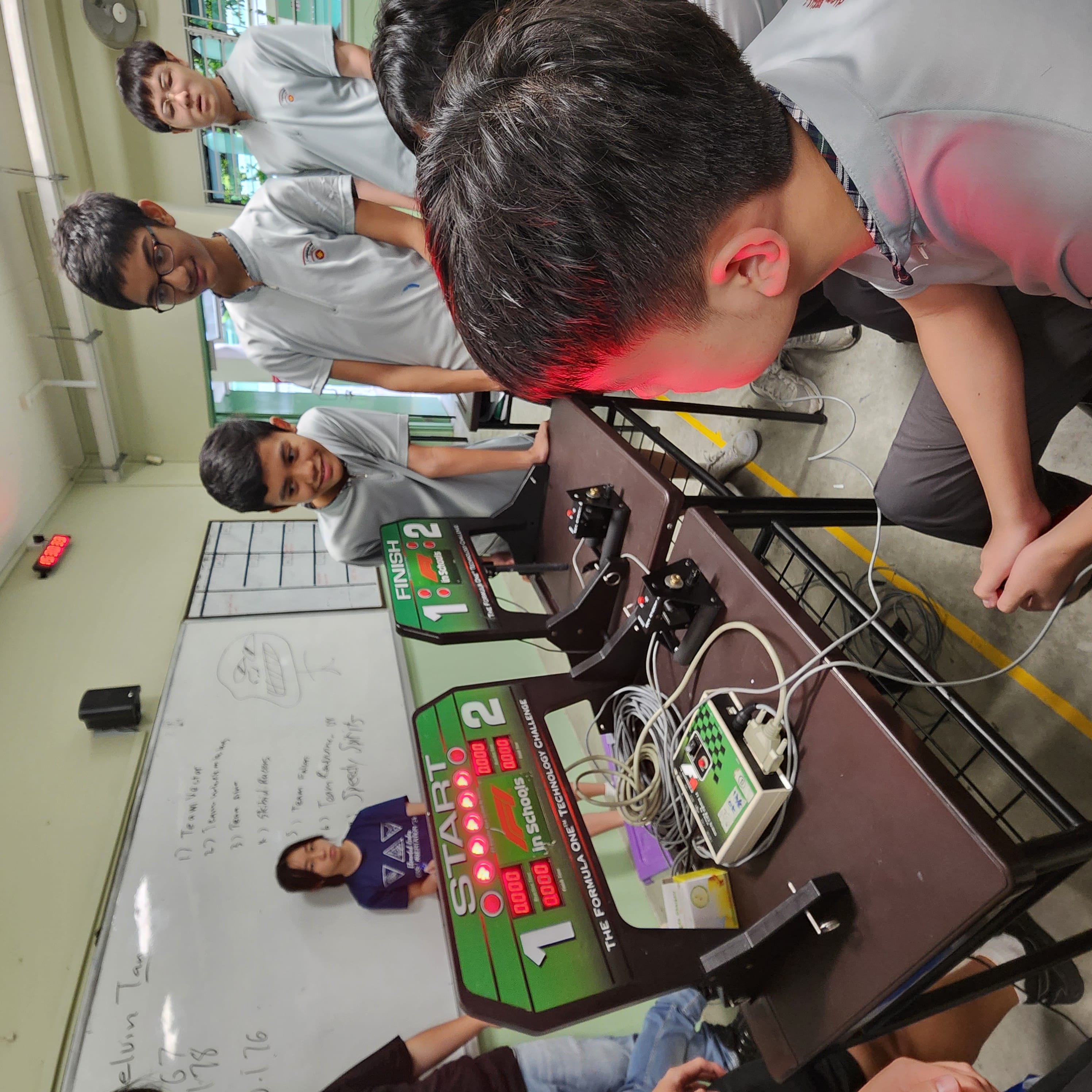Students gather around a Formula One in Schools Technology Challenge start and finish line timing system in a classroom.