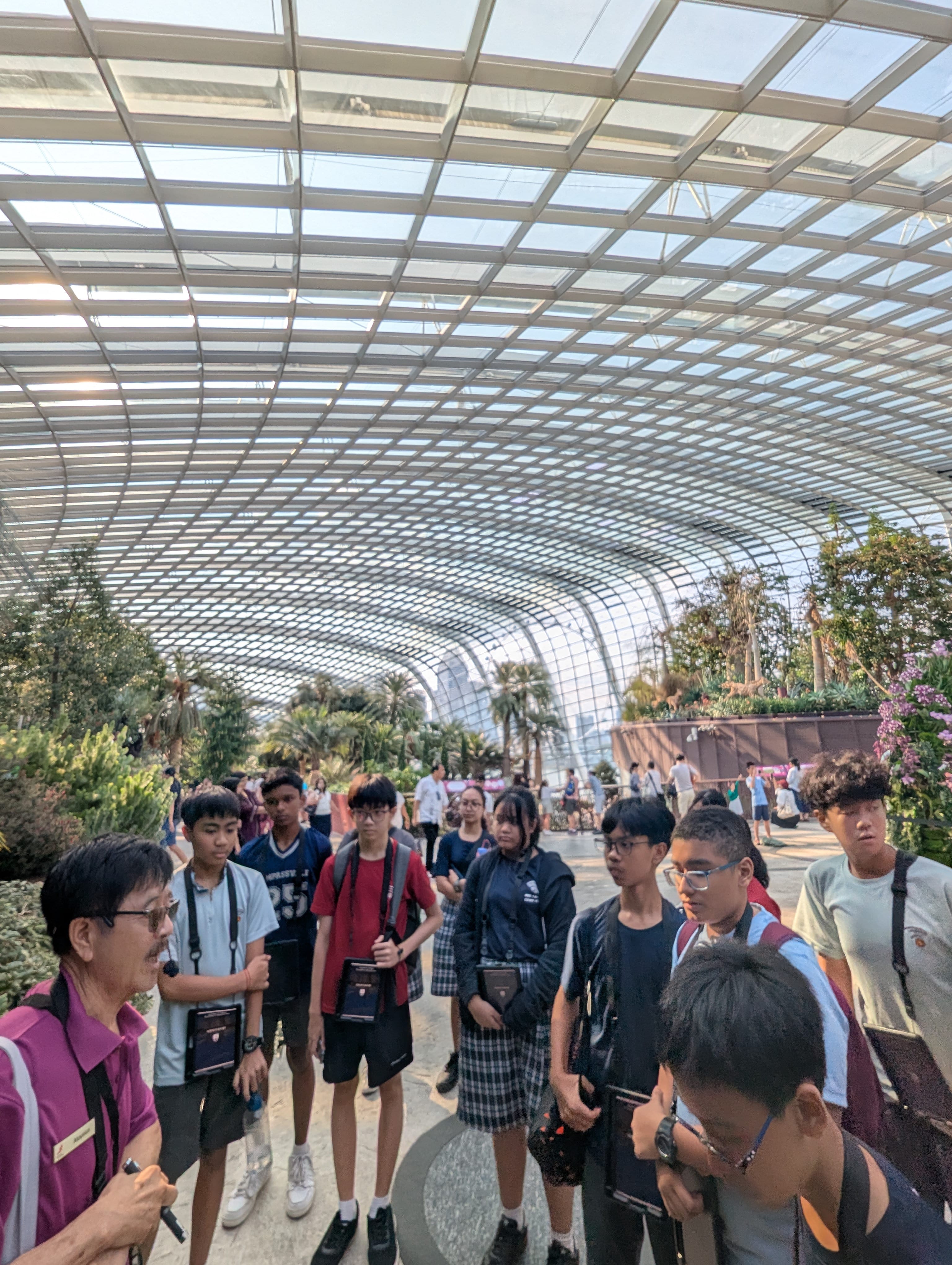 A guide in a pink shirt leads a group of teenagers inside a large glass-domed greenhouse with lush plants.