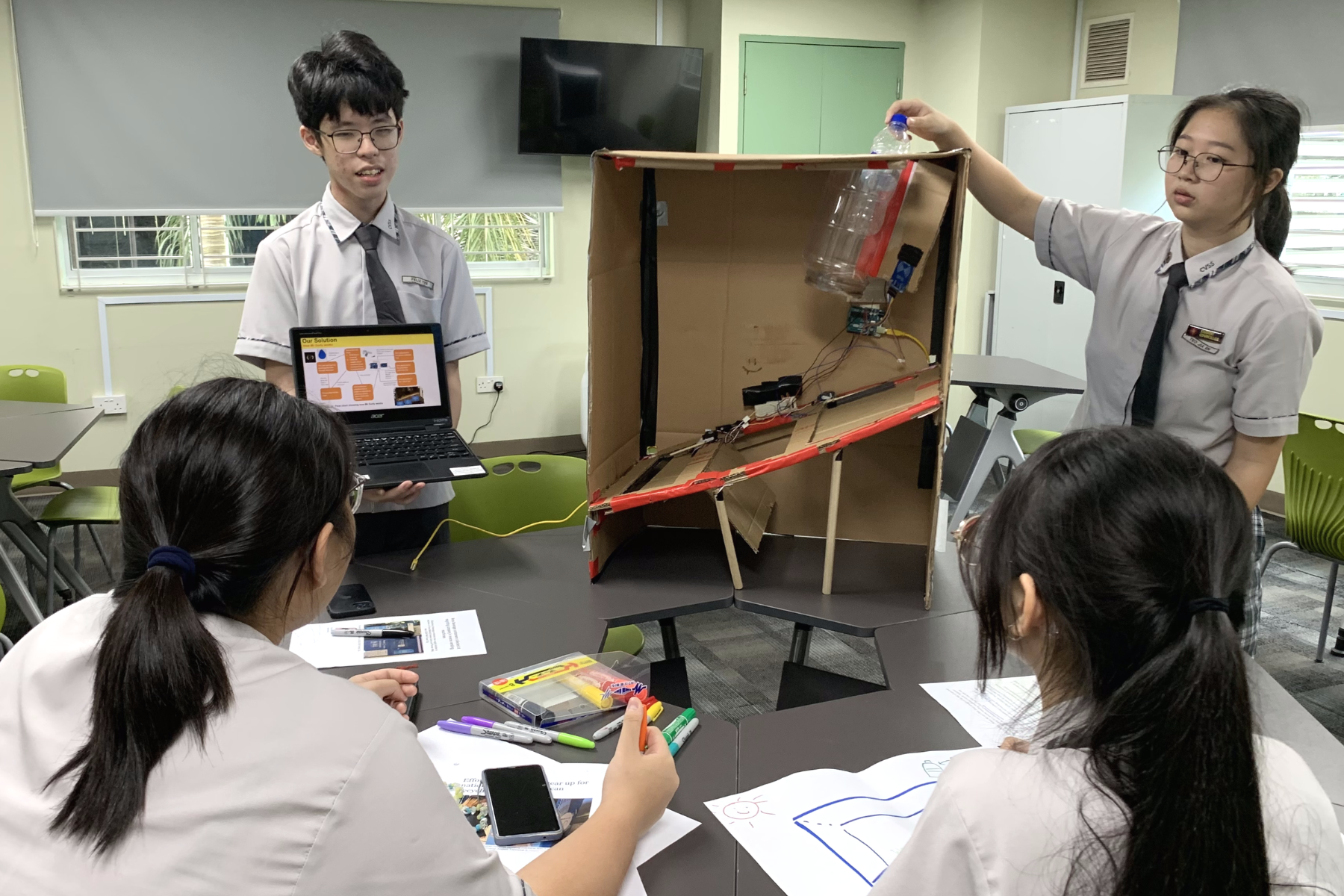 Four students in school uniforms present a cardboard prototype with electronics to seated classmates in a classroom.