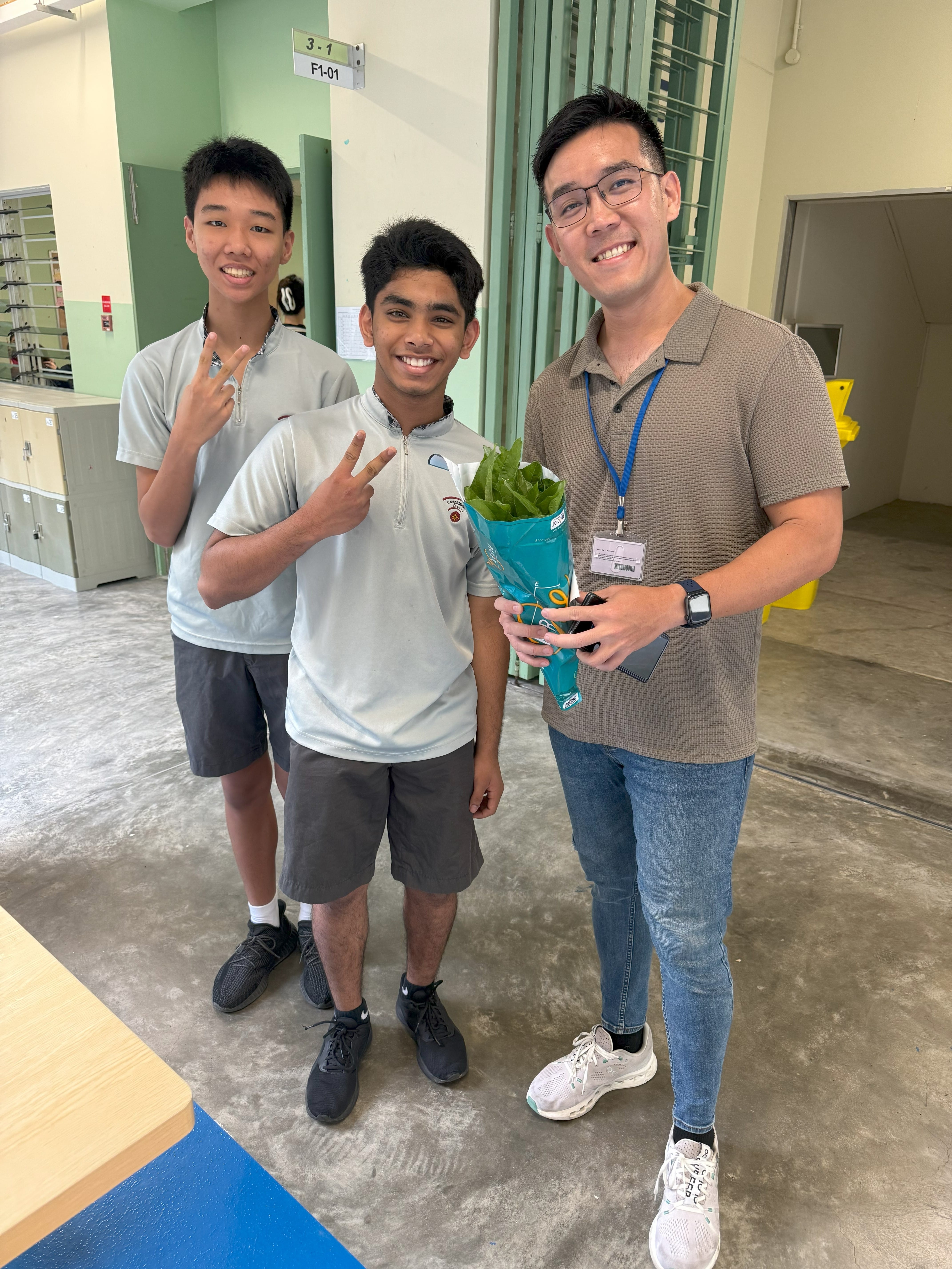 Three smiling people indoors; two teens flash peace signs while an adult with glasses holds a leafy bouquet.