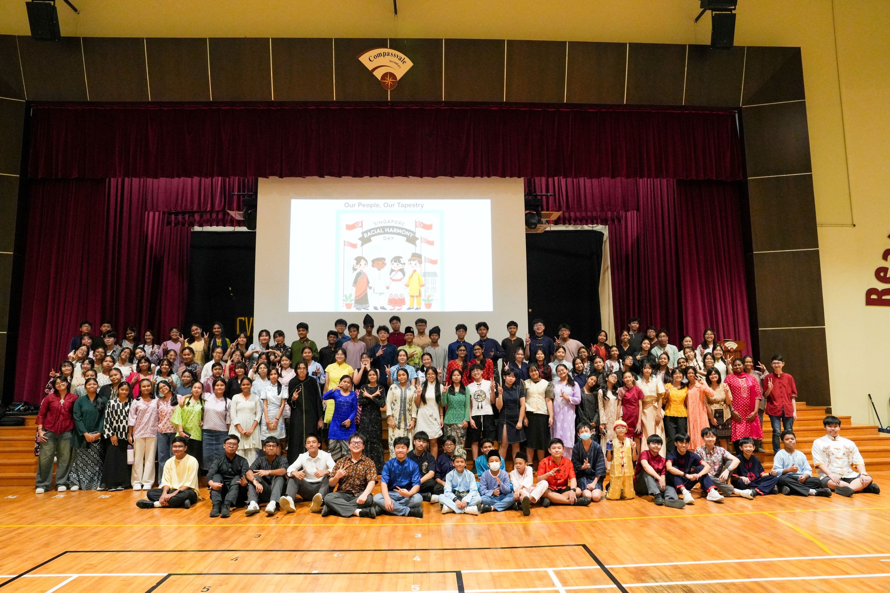 Large group of students in diverse traditional costumes pose in a school hall for Singapore Racial Harmony Day.