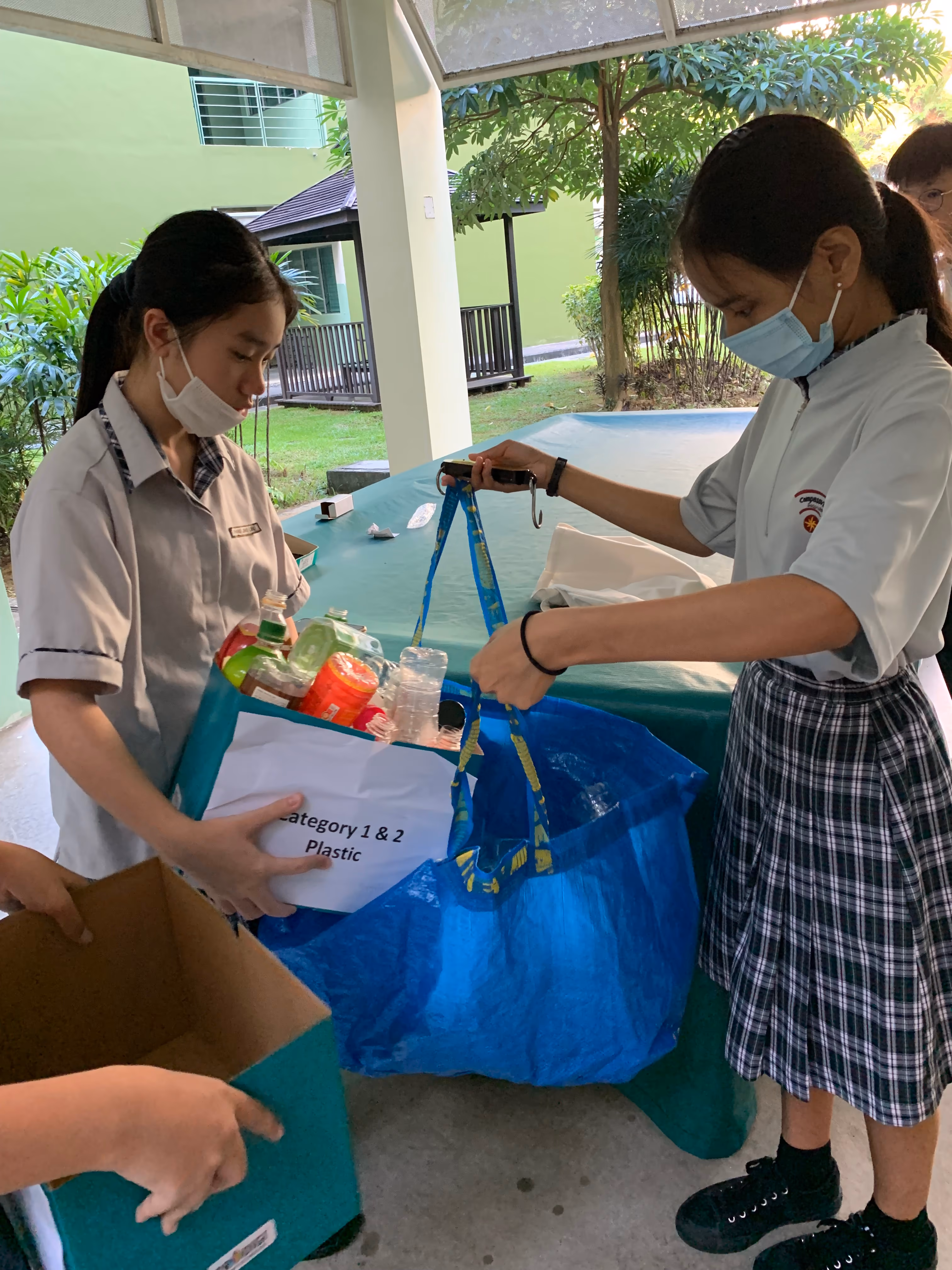 Two masked students in uniforms sort plastic bottles into a blue bag labeled Category 1 and 2 Plastic outdoors.