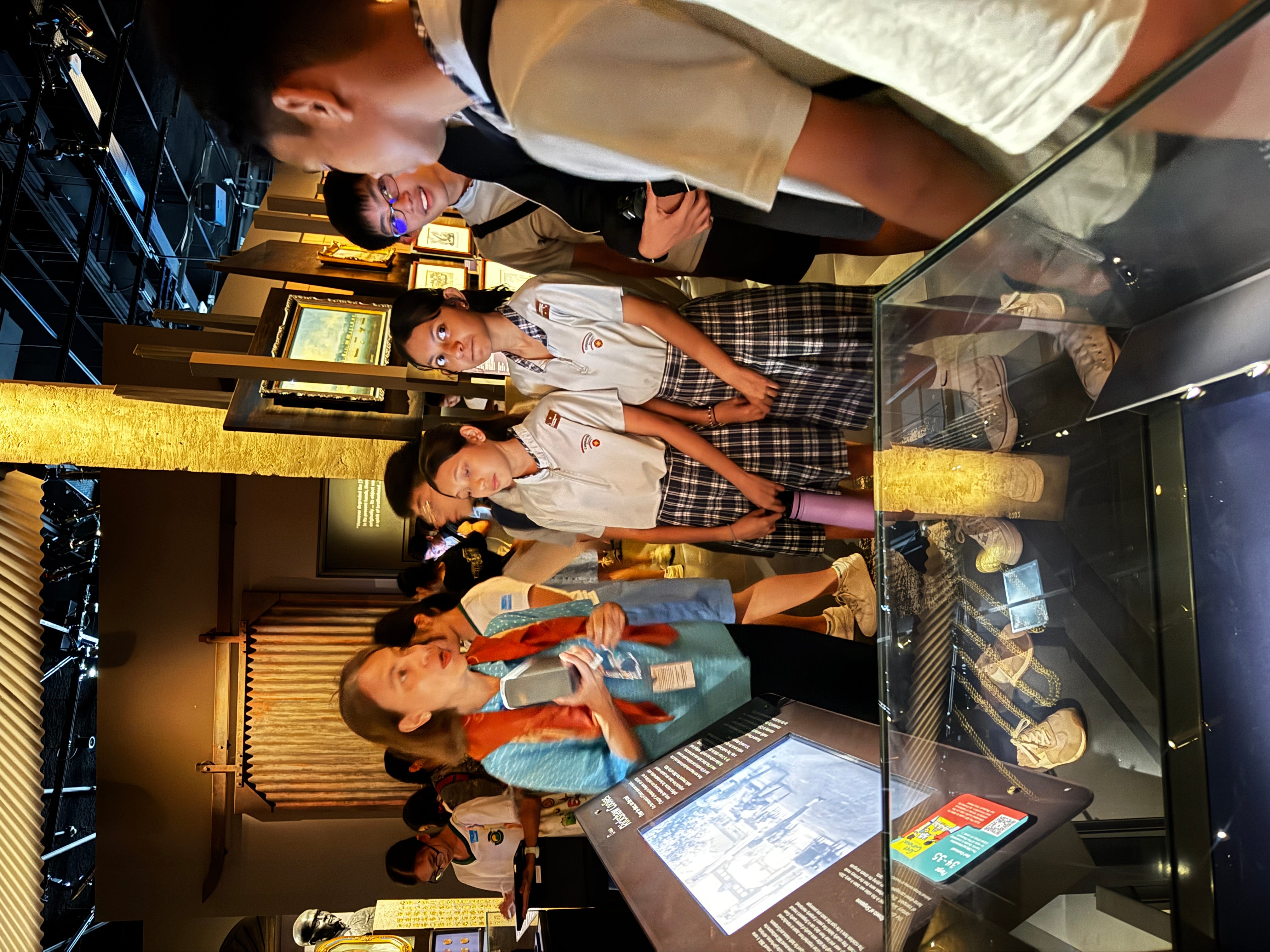 A museum guide speaks to students in plaid uniforms gathered around a display case in a dimly lit exhibit hall.
