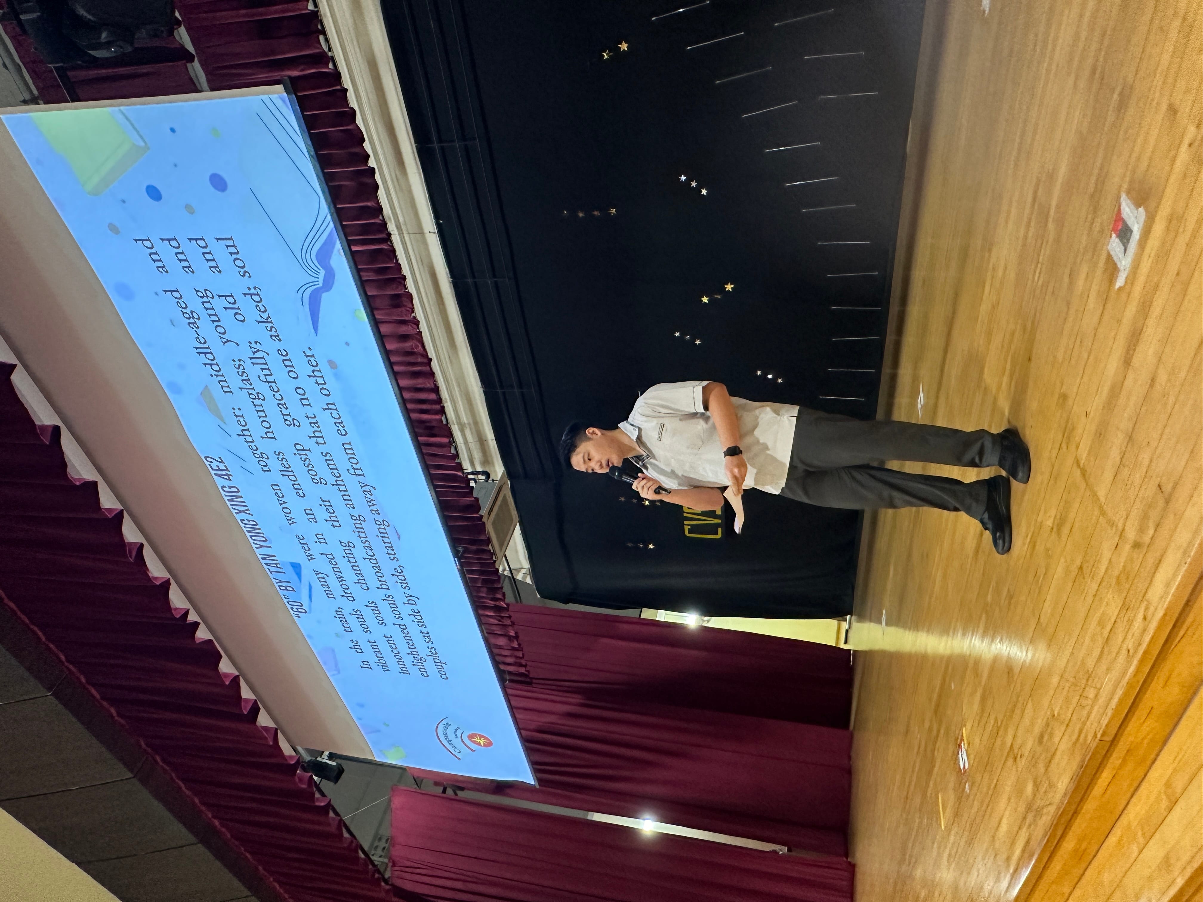A man holds a microphone on a school stage with a projected slide showing a poem titled "60" by Tan Yong Xing 4E2.
