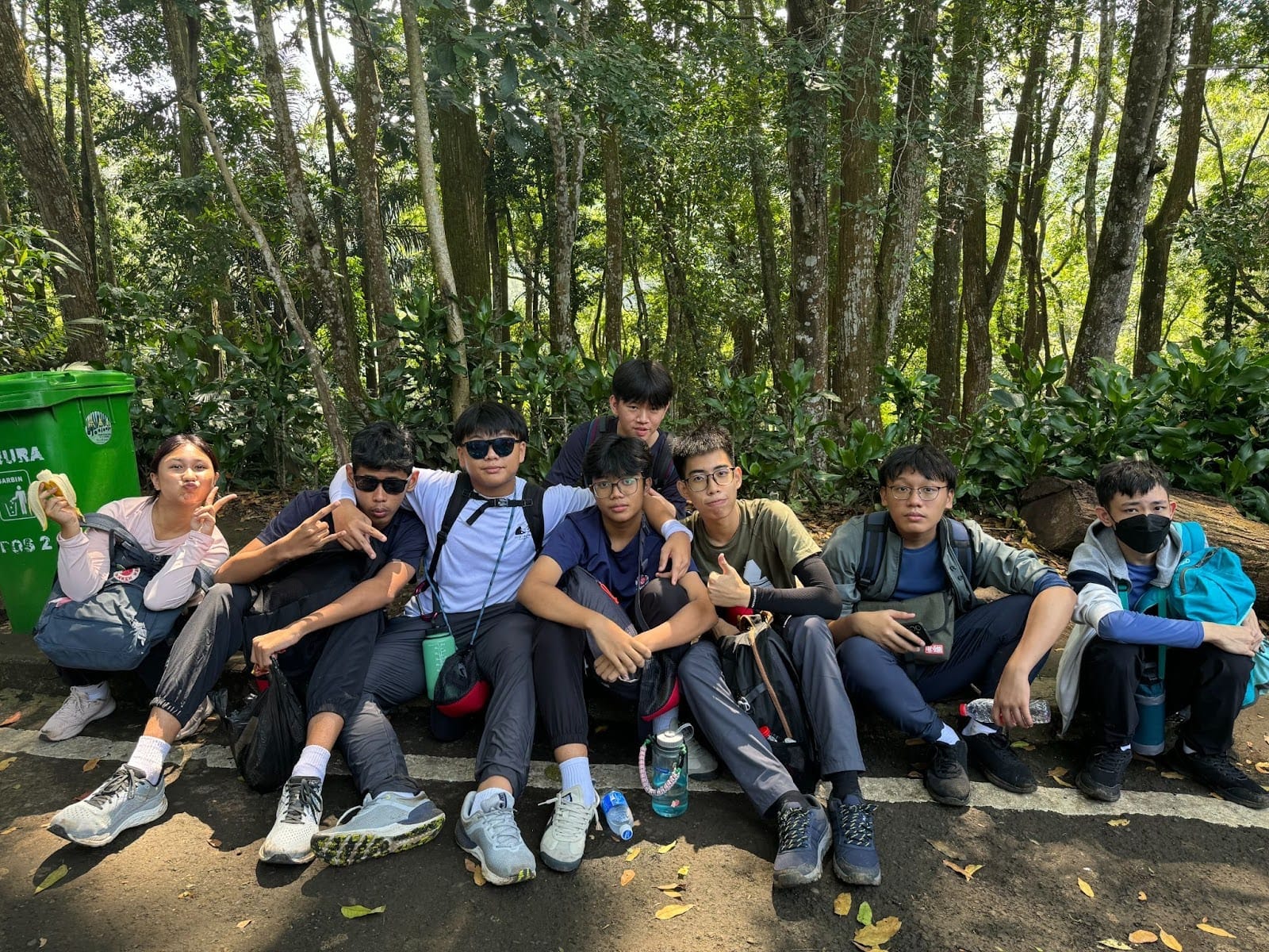 A group of about nine young teens sitting and posing together on the ground in a forested area with tall trees.