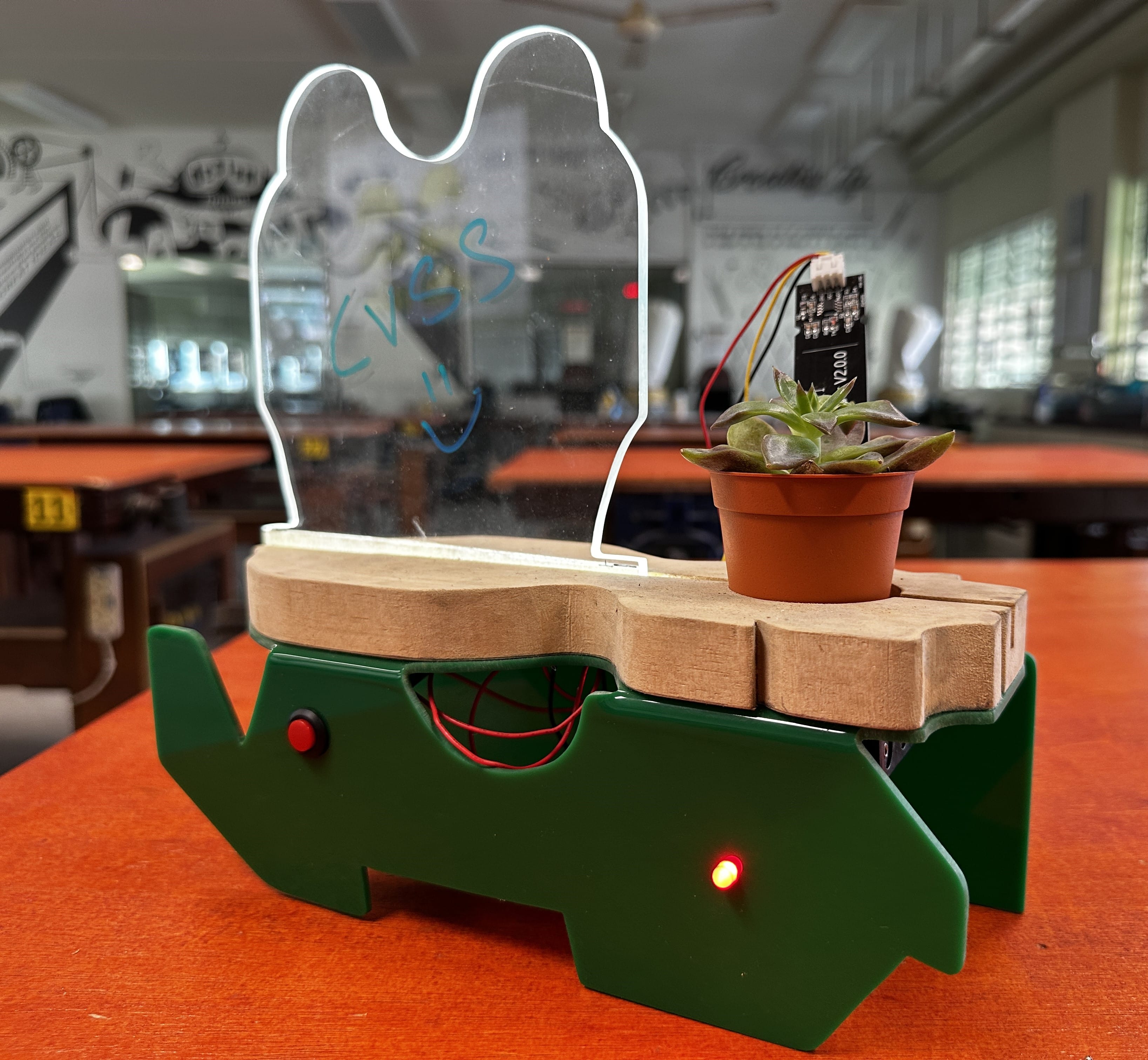 A green robot-shaped device with a wooden top, small succulent plant, acrylic display, and LED lights on a red table.