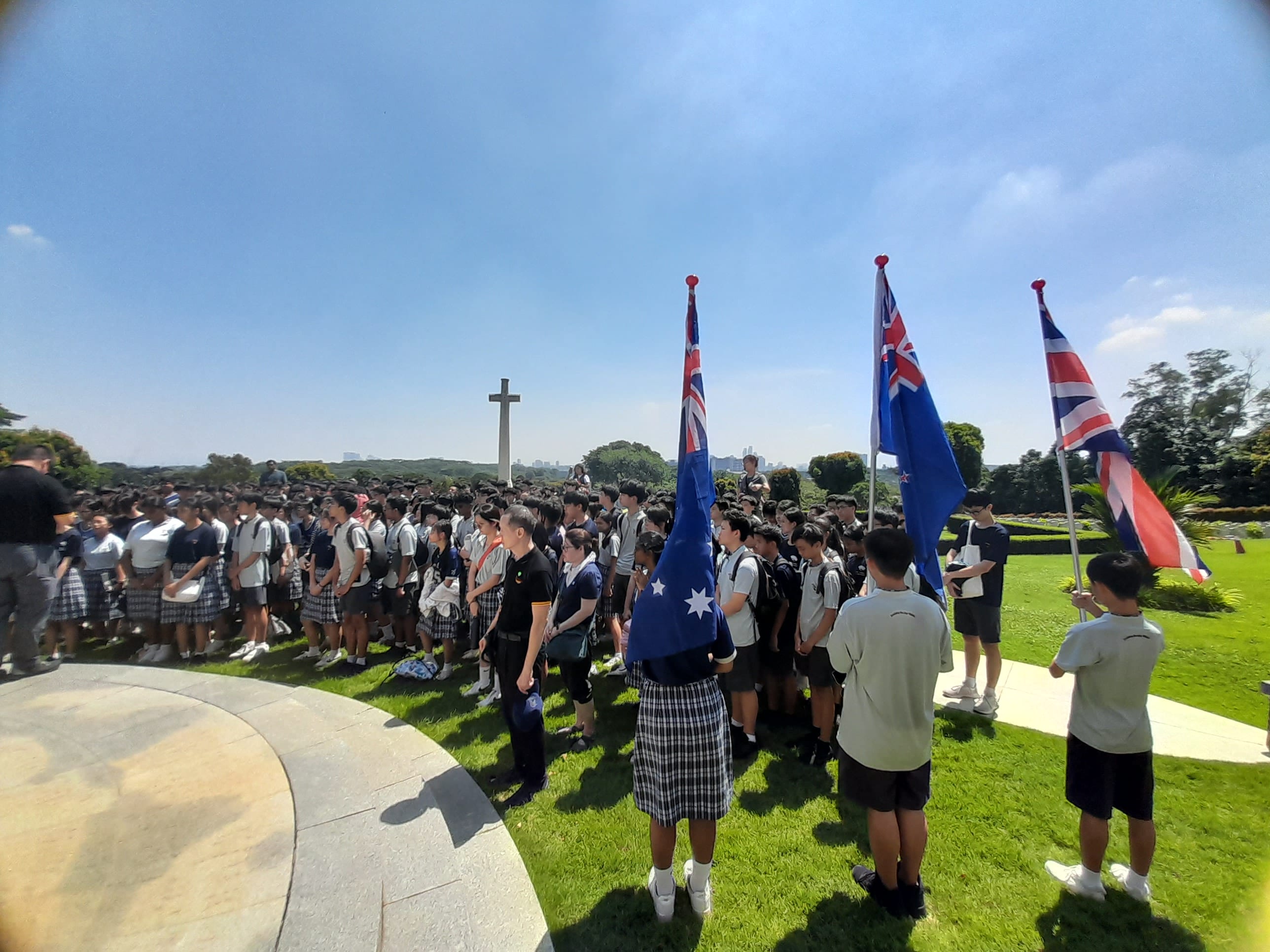 Students in uniforms gather outdoors near a stone cross monument, with Australian, New Zealand, and UK flags being held.