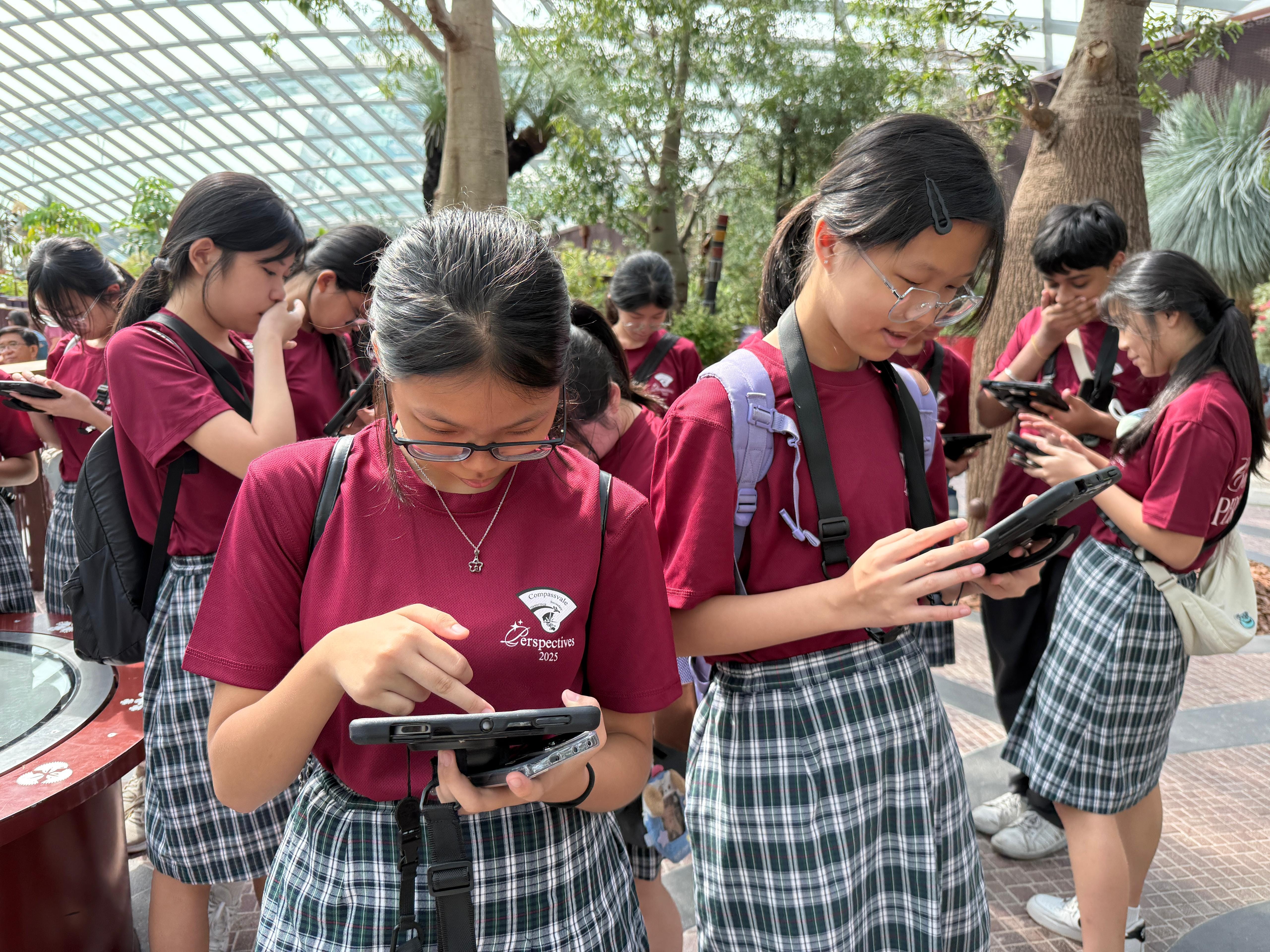 A group of students in maroon shirts and plaid skirts look at phones and tablets inside a glass-roofed greenhouse.
