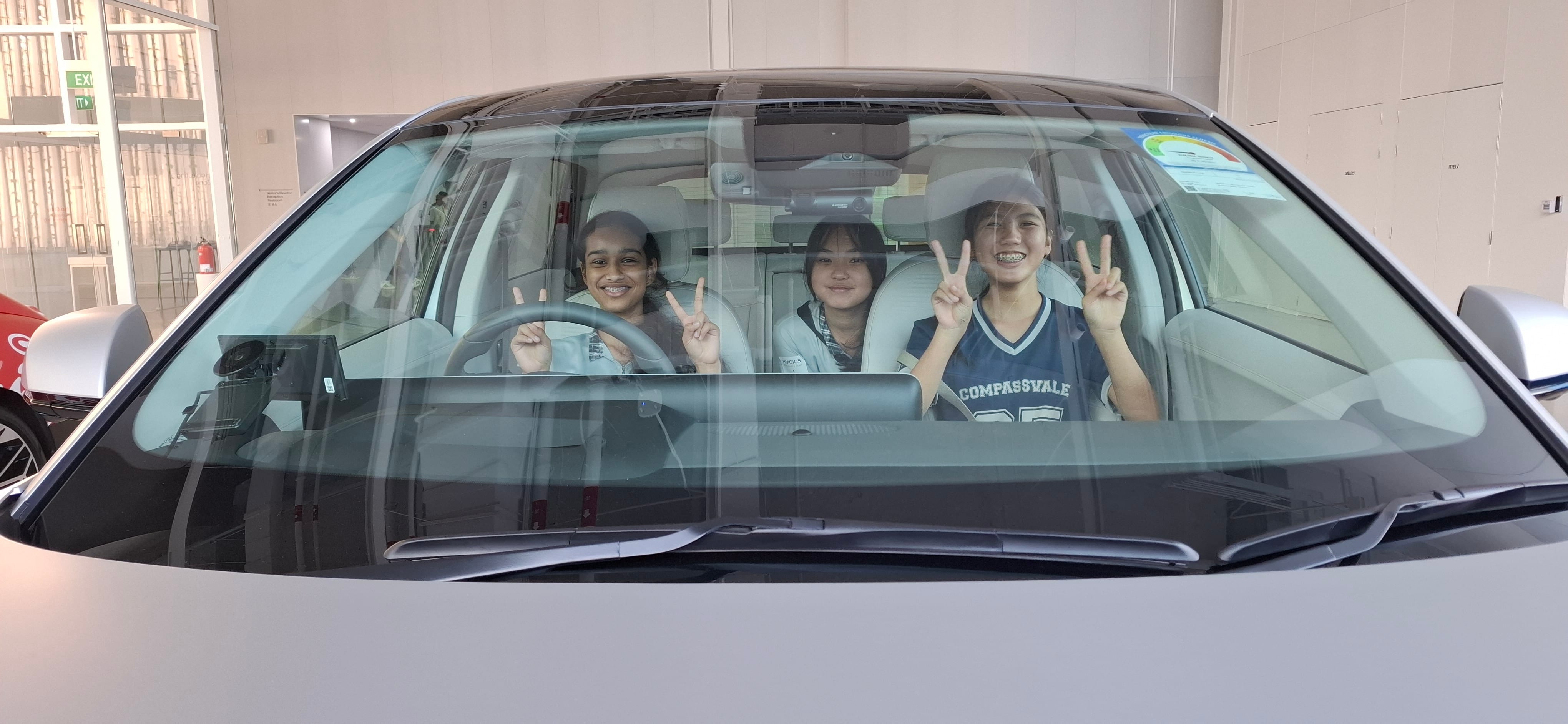 Three smiling girls sit inside a car, visible through the windshield, making peace signs with their hands.