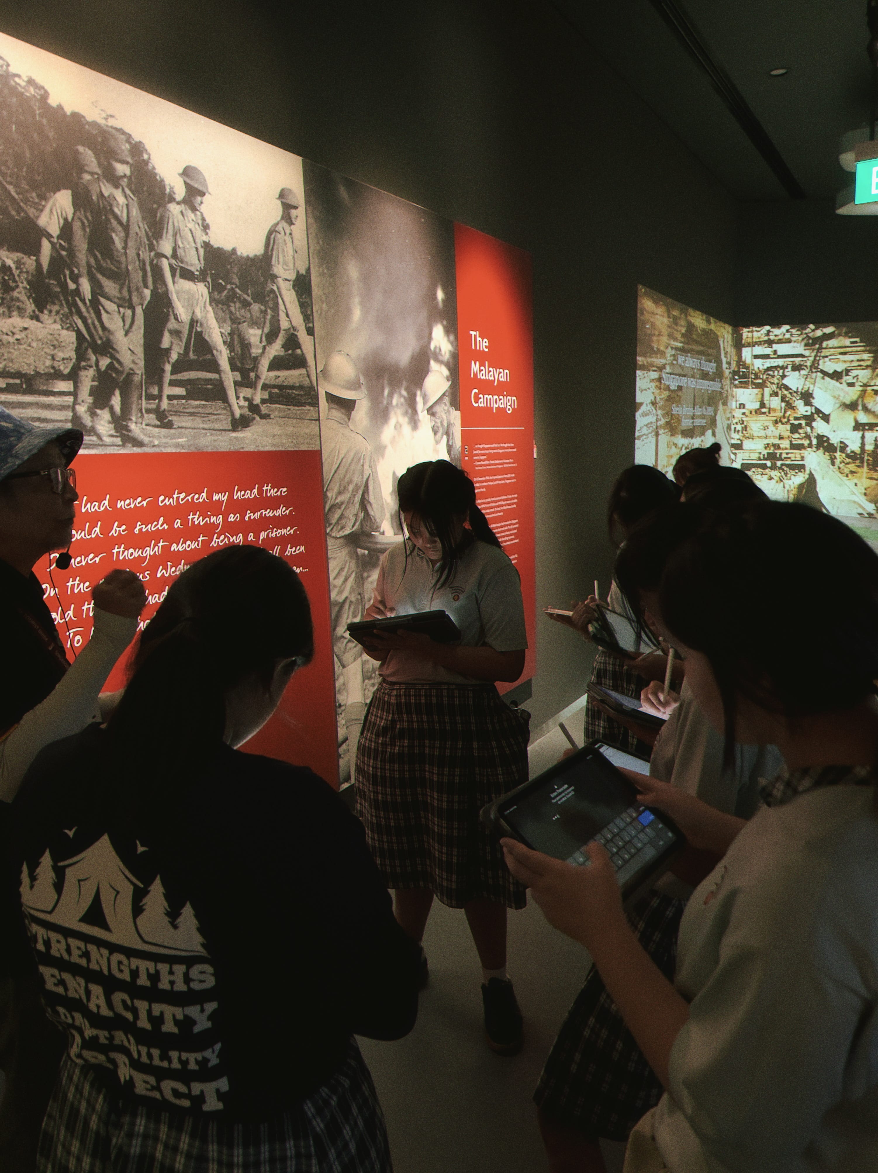 Students with tablets visit a museum exhibit titled The Malayan Campaign, featuring wartime photos and red display panels.