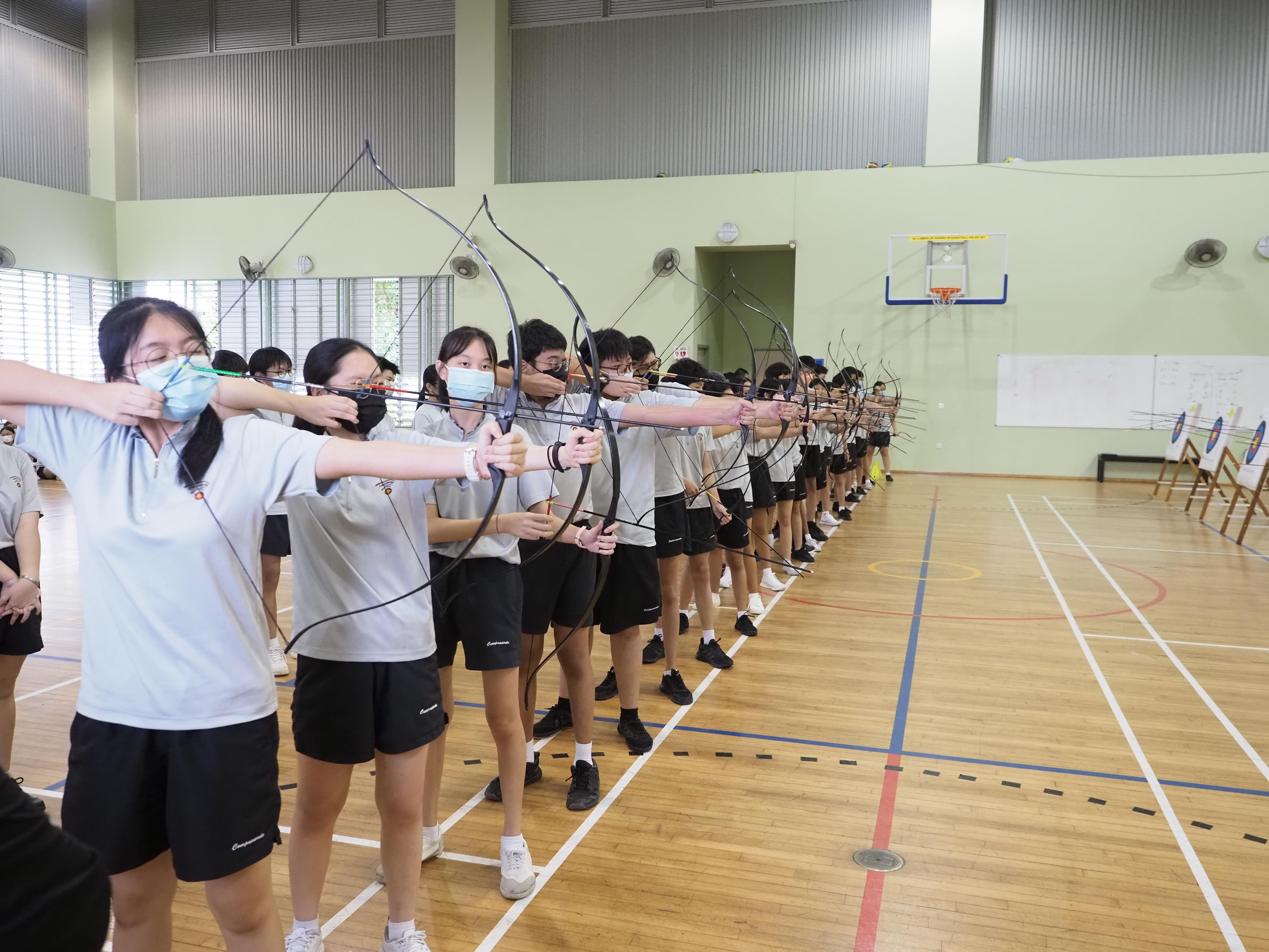 A row of students in white shirts aim bows and arrows toward targets in an indoor gymnasium.
