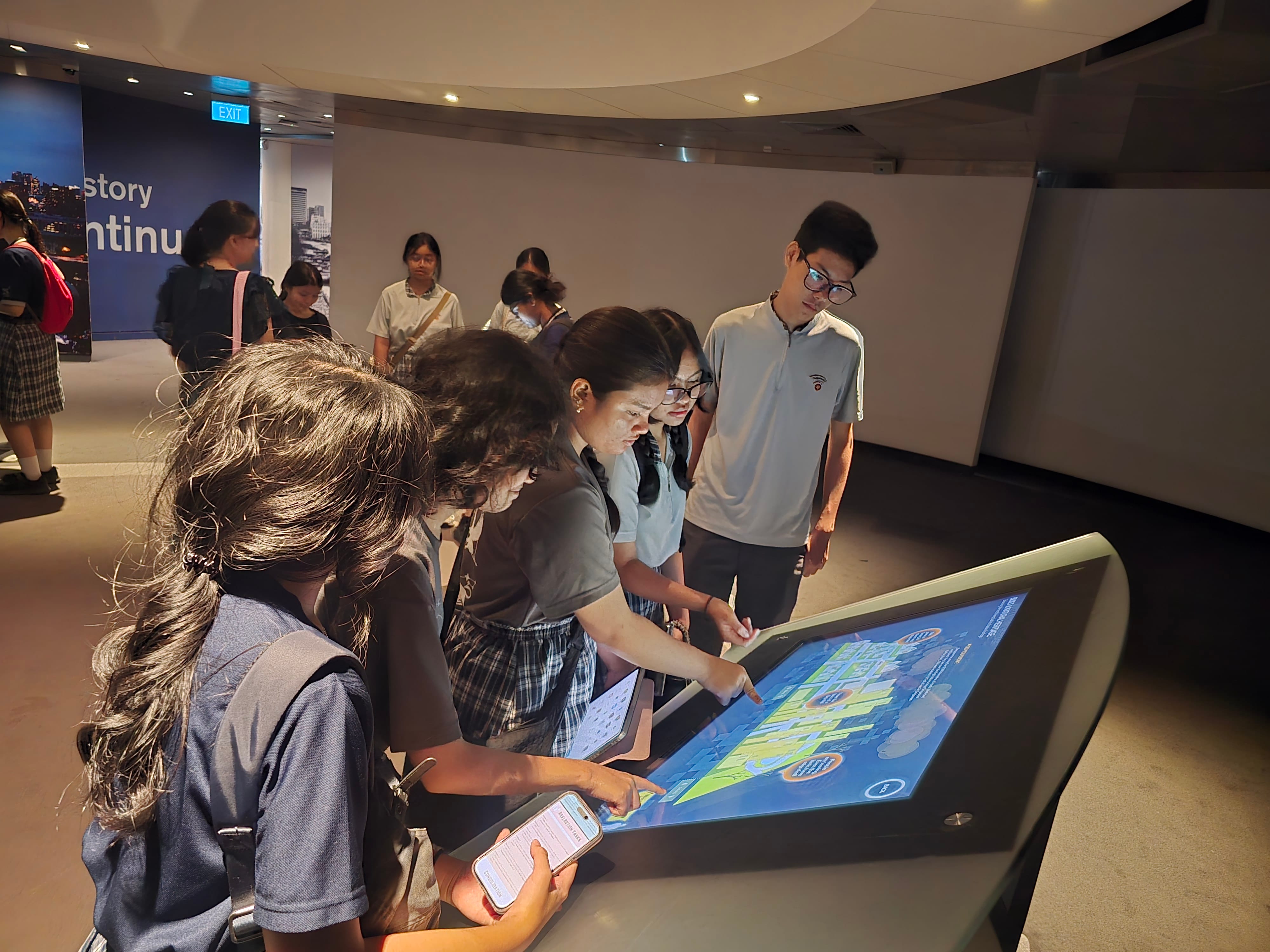 Students in school uniforms gather around a large interactive touchscreen display in a dimly lit museum room.