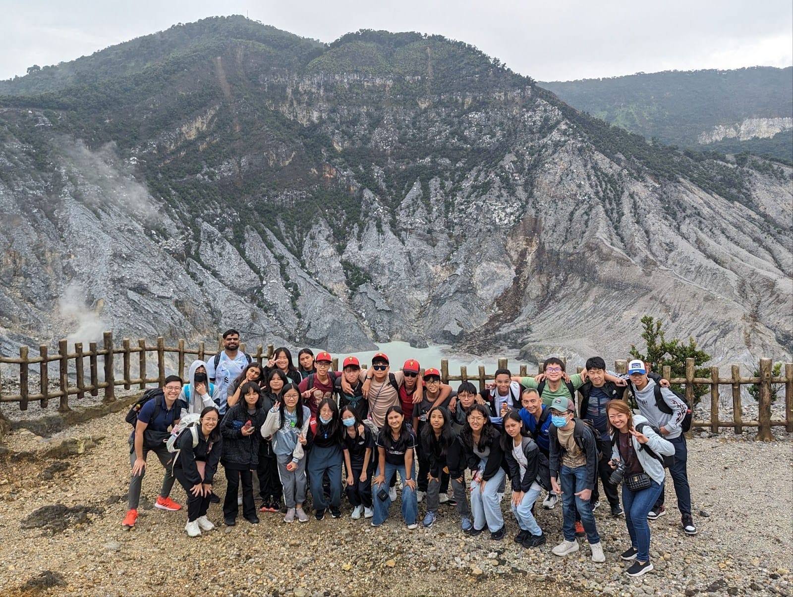 A large group of people posing in front of a volcanic crater with steam vents and rocky mountains behind them.