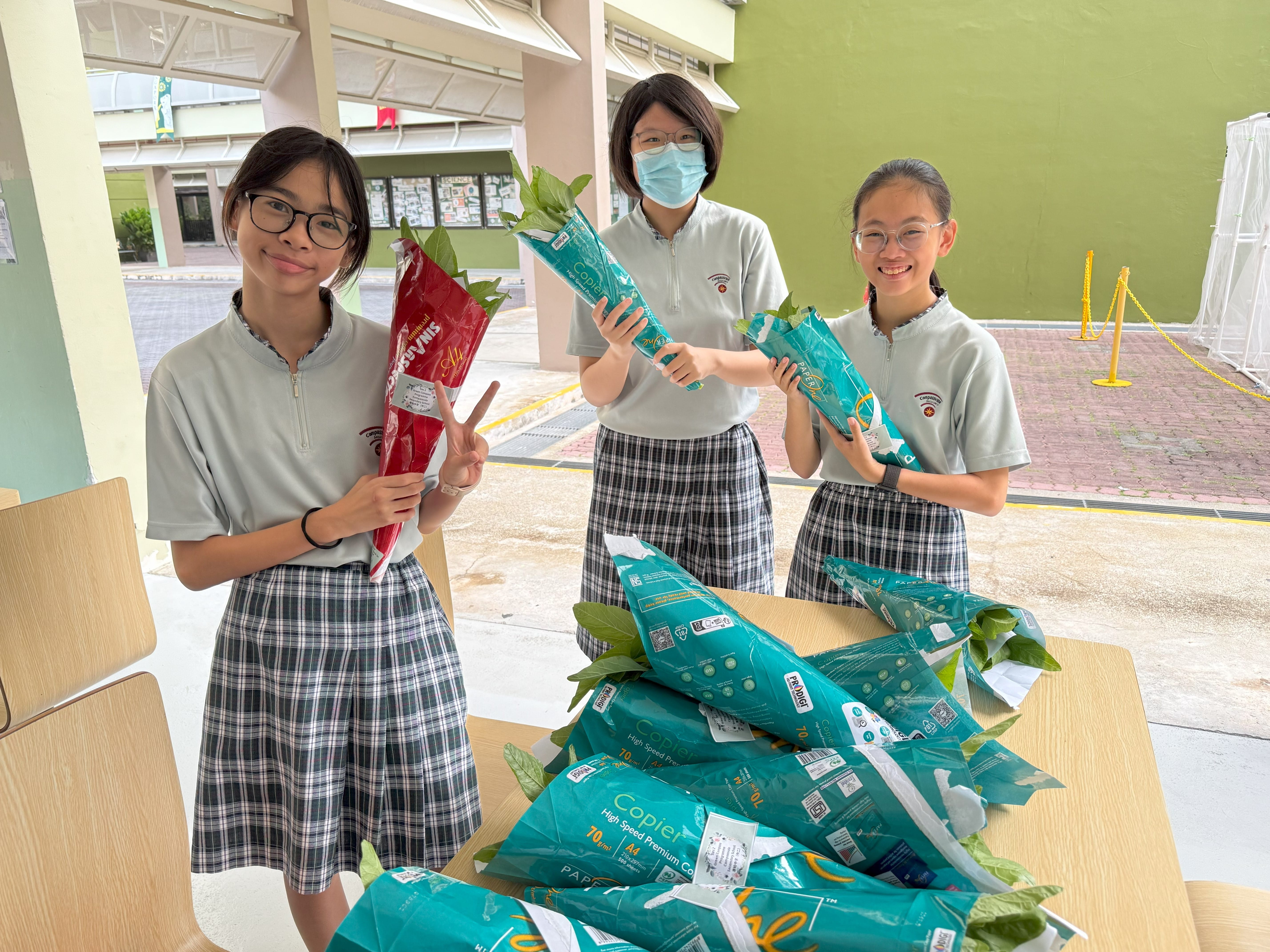 Three students in school uniforms hold wrapped plants by a table piled with more wrapped plants.