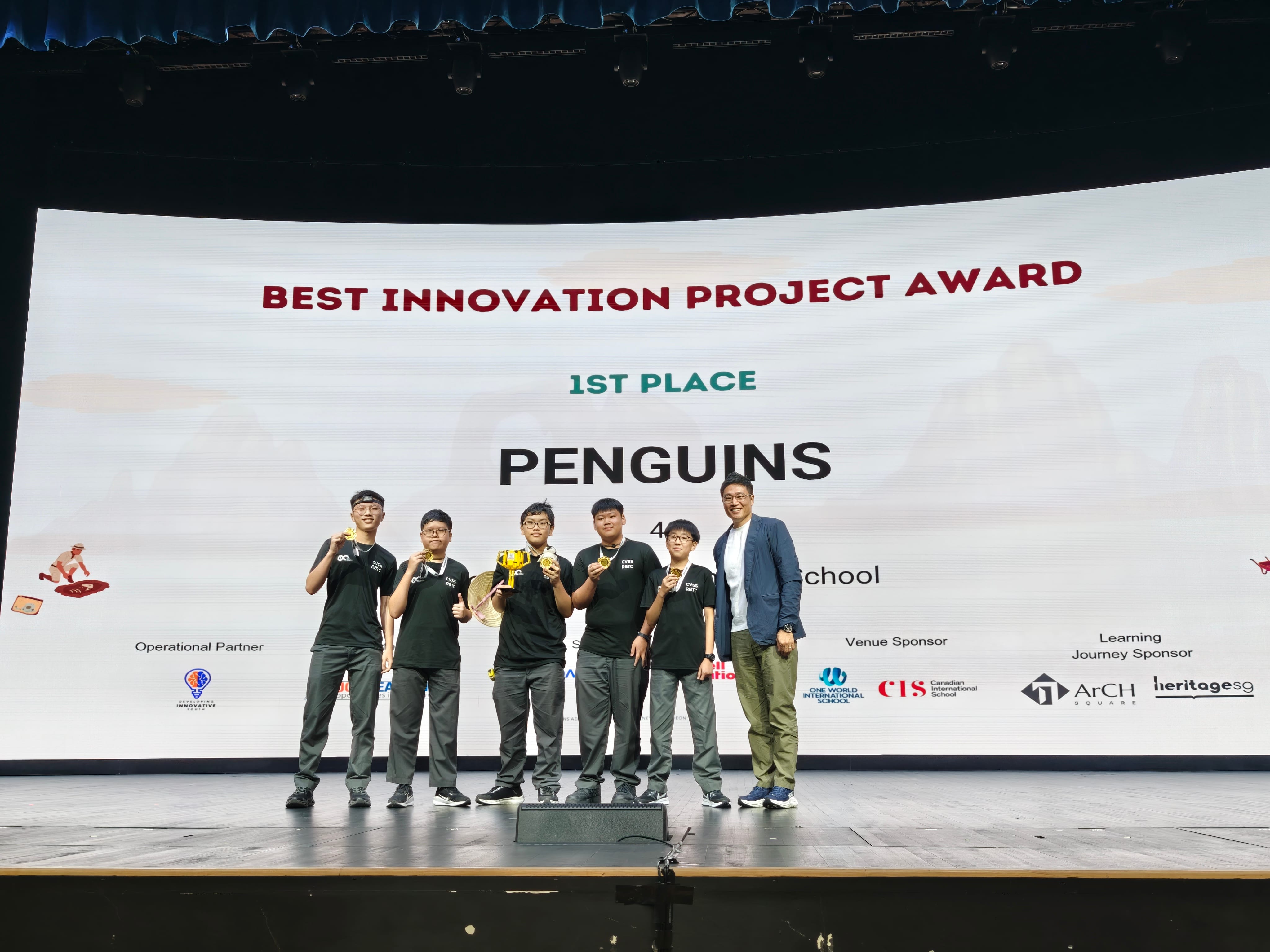 Five students and an adult on stage holding a trophy, in front of a screen showing Best Innovation Project Award 1st Place Penguins.