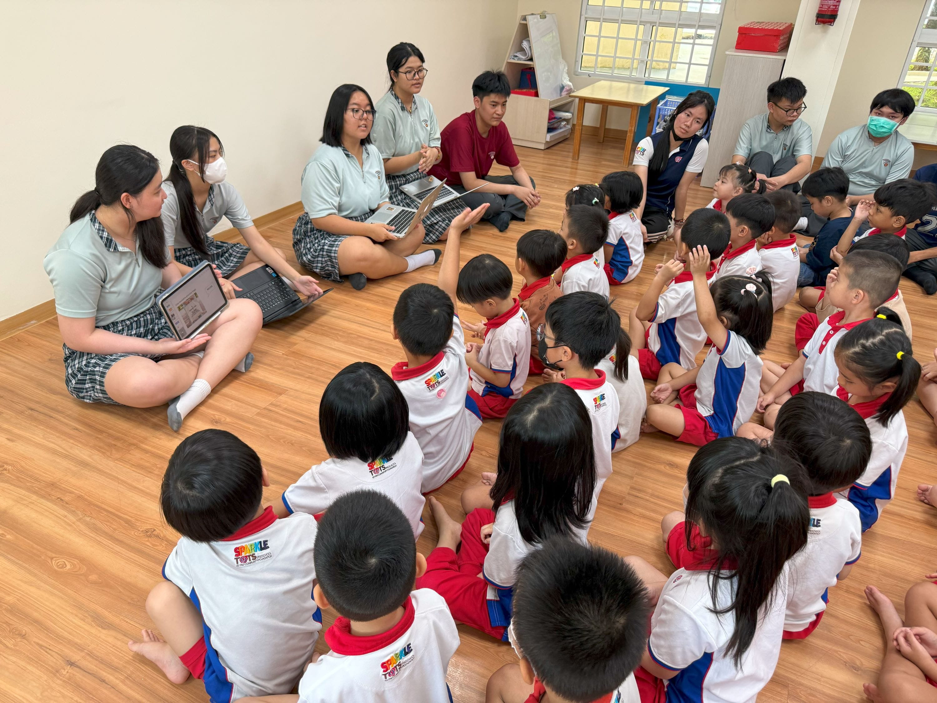 Teenagers with laptops sit on a wooden floor facing a large group of young children in red and white uniforms.