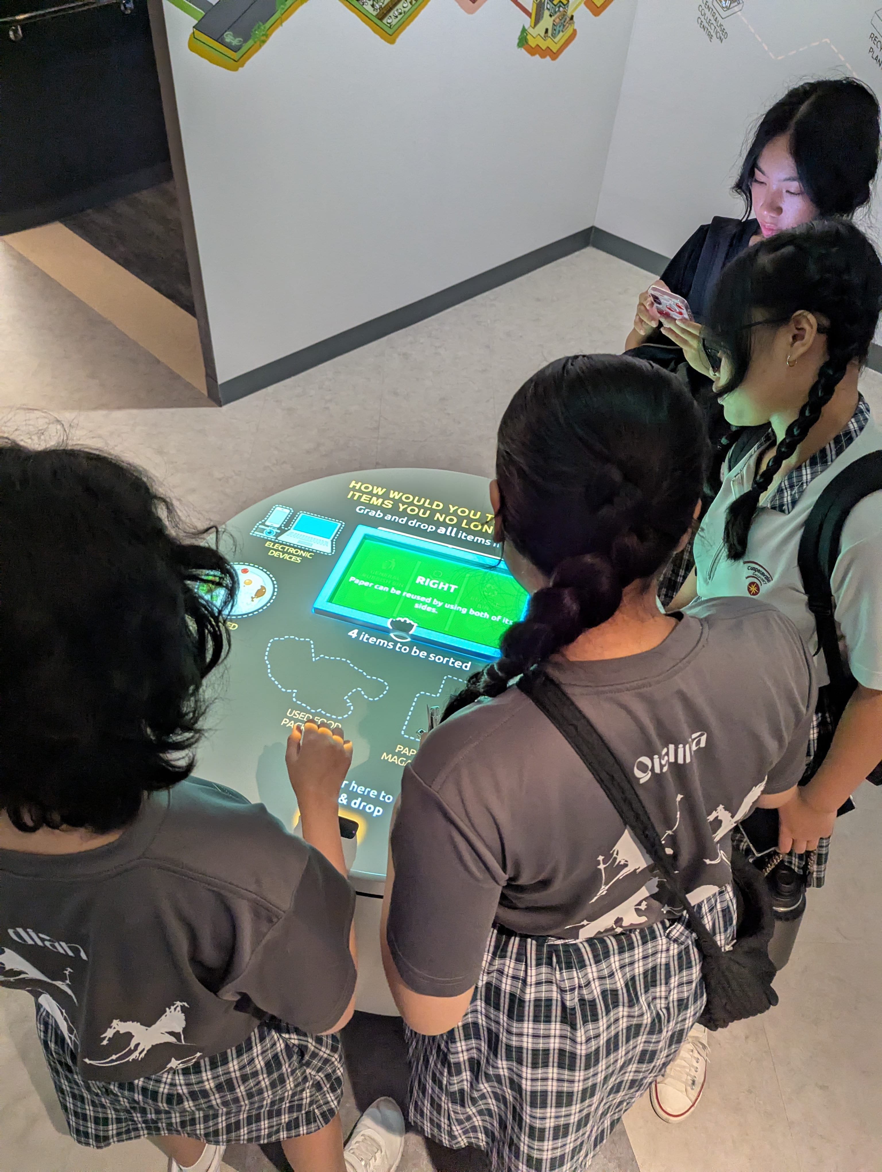 Students in school uniforms gather around an interactive touchscreen table displaying a recycling sorting activity at a museum.