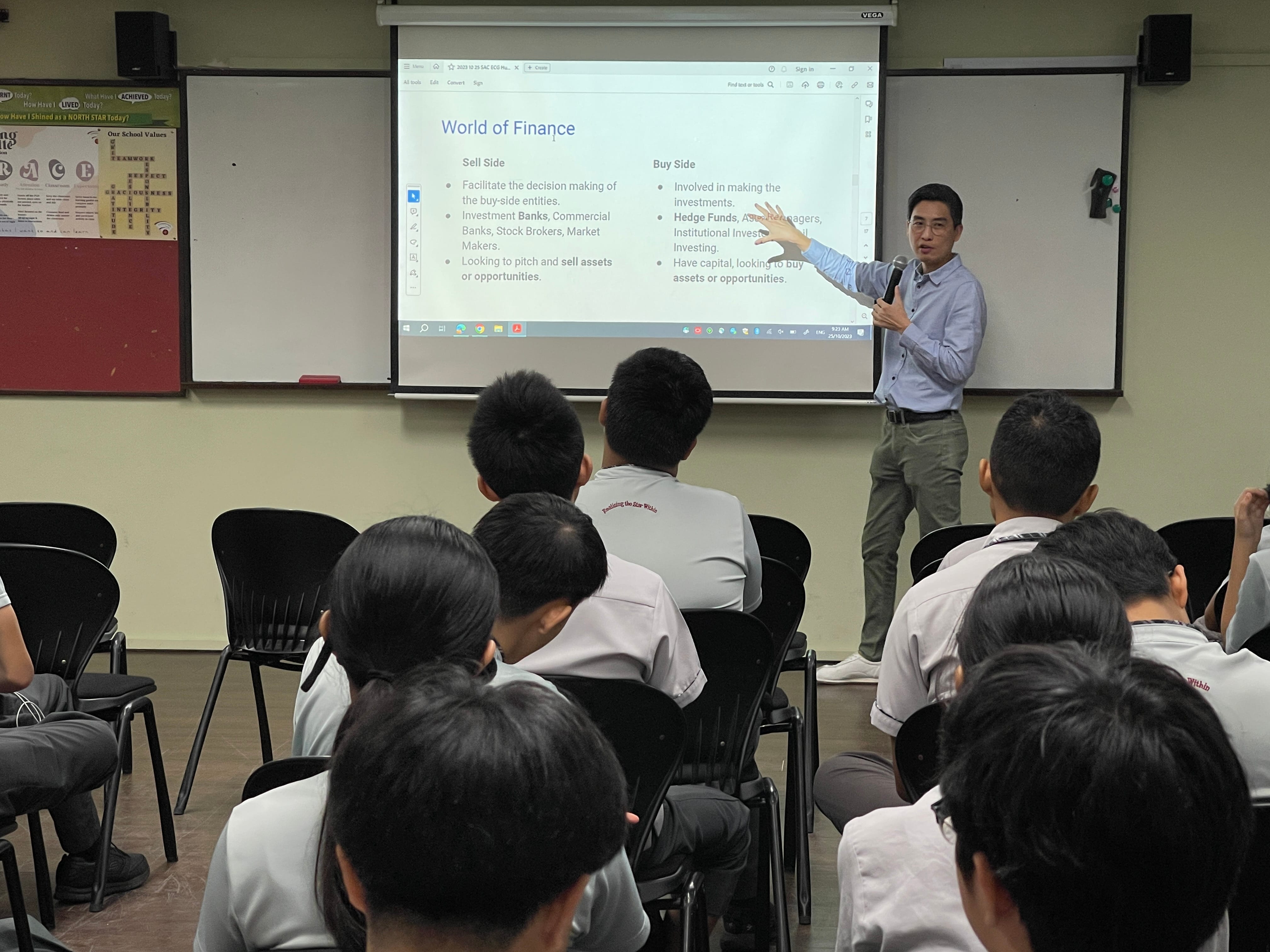 A man presents a World of Finance slideshow to students in school uniforms seated in a classroom.