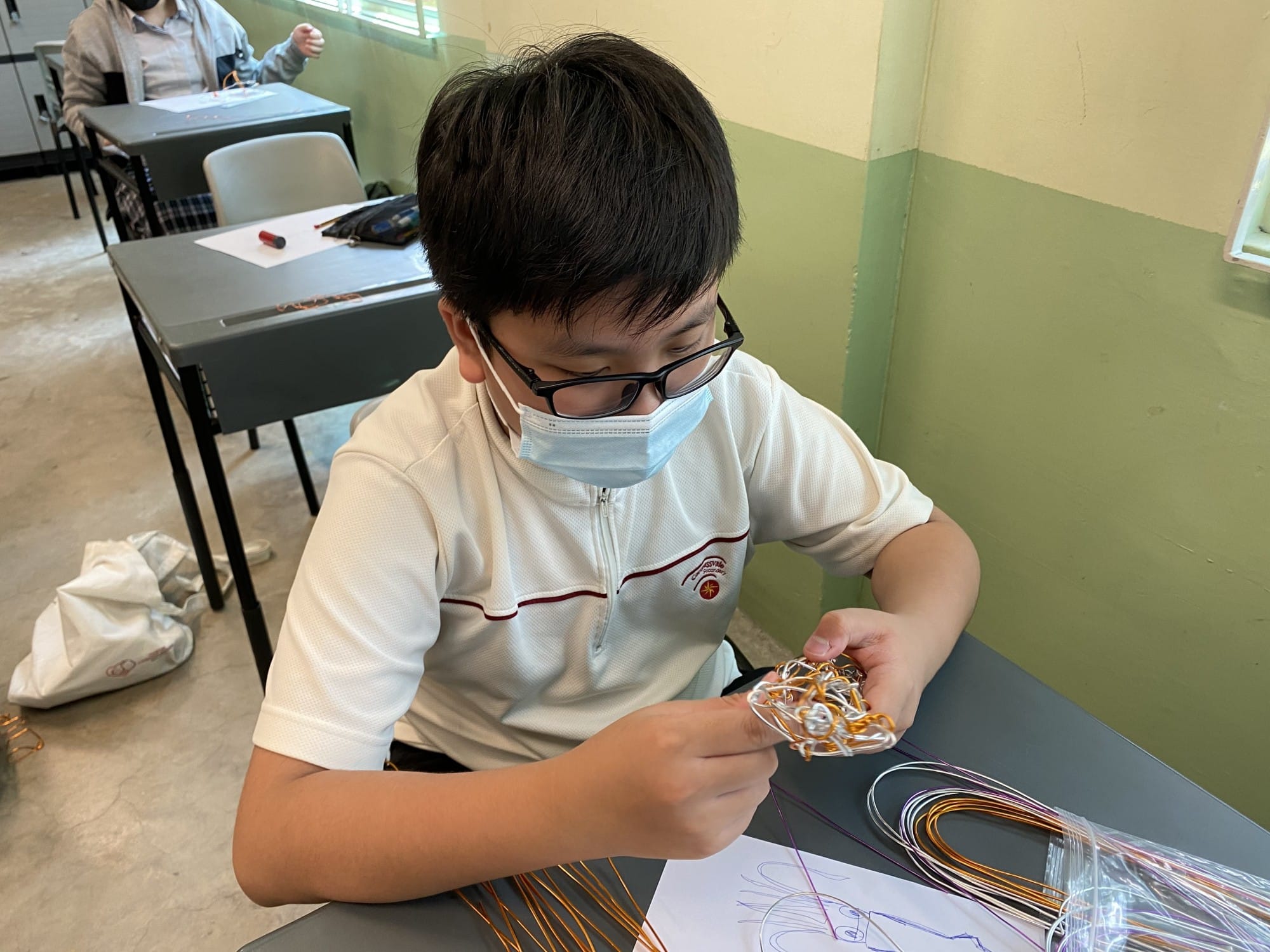 A boy wearing glasses and a face mask sits at a desk, carefully bending colorful wires into a sculpture.