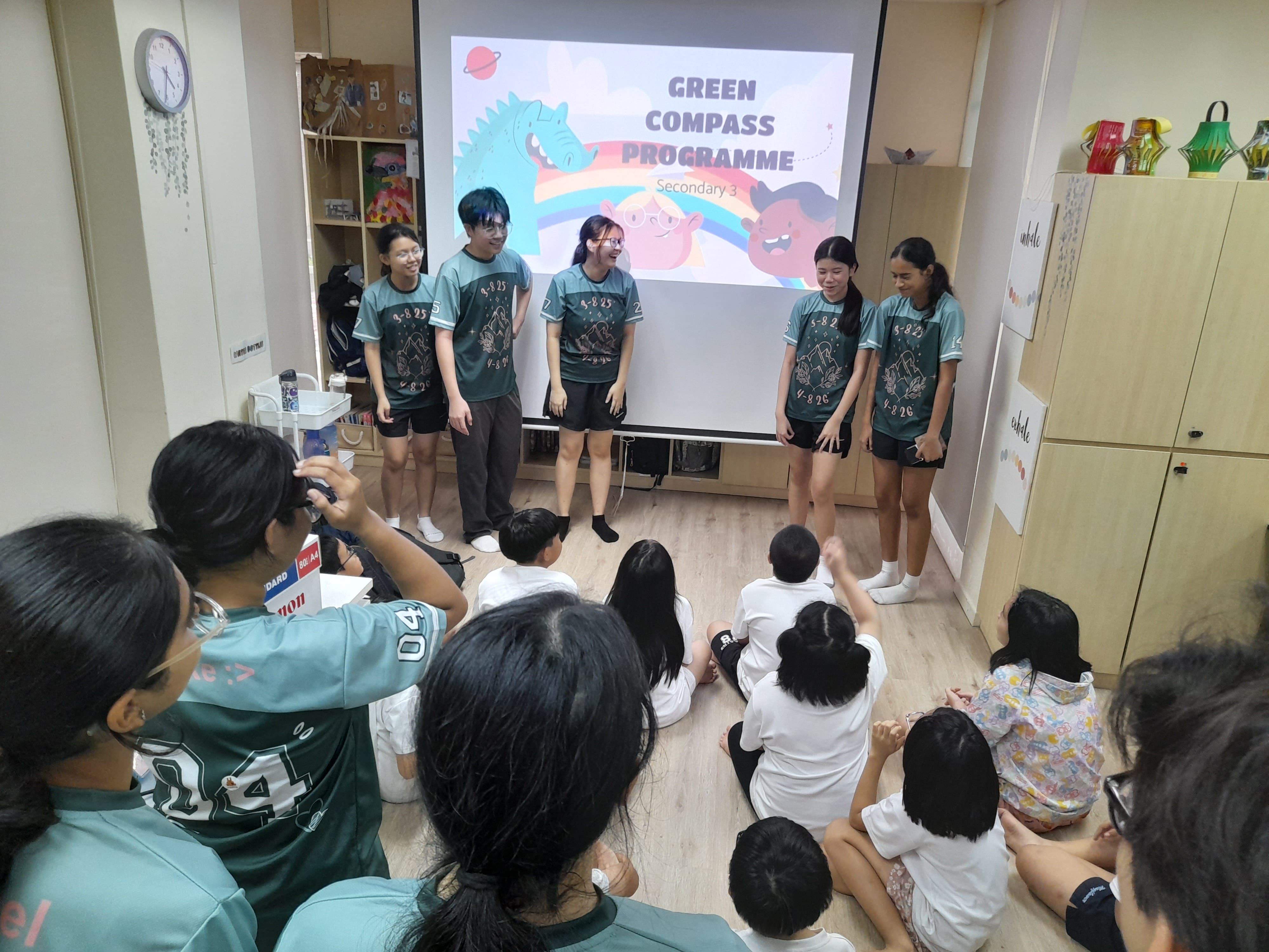 Five students in green shirts present the Green Compass Programme to younger students seated on the floor.