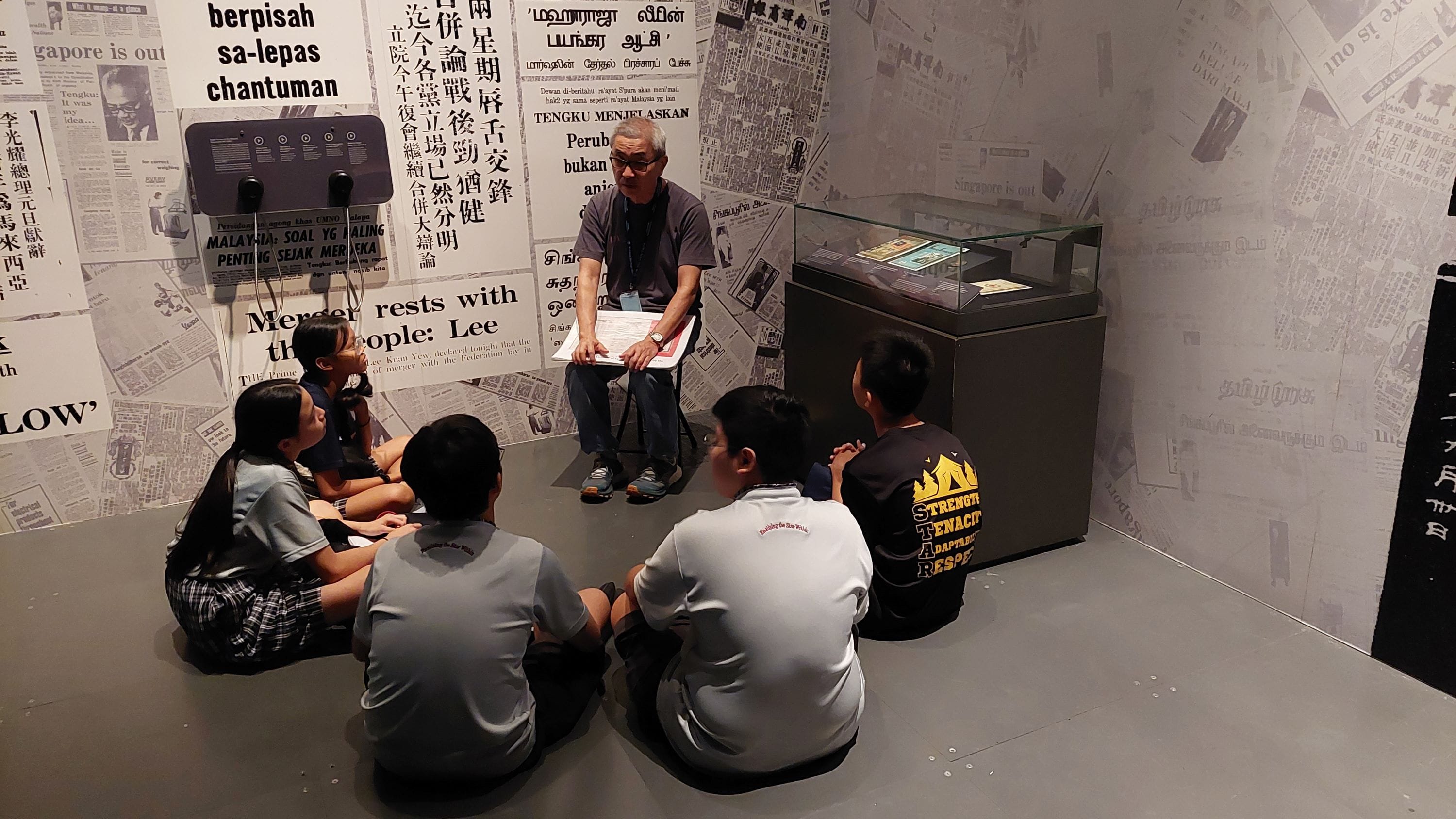 An adult guide speaks to children seated on a floor in a museum room with multilingual newspaper clippings on walls.