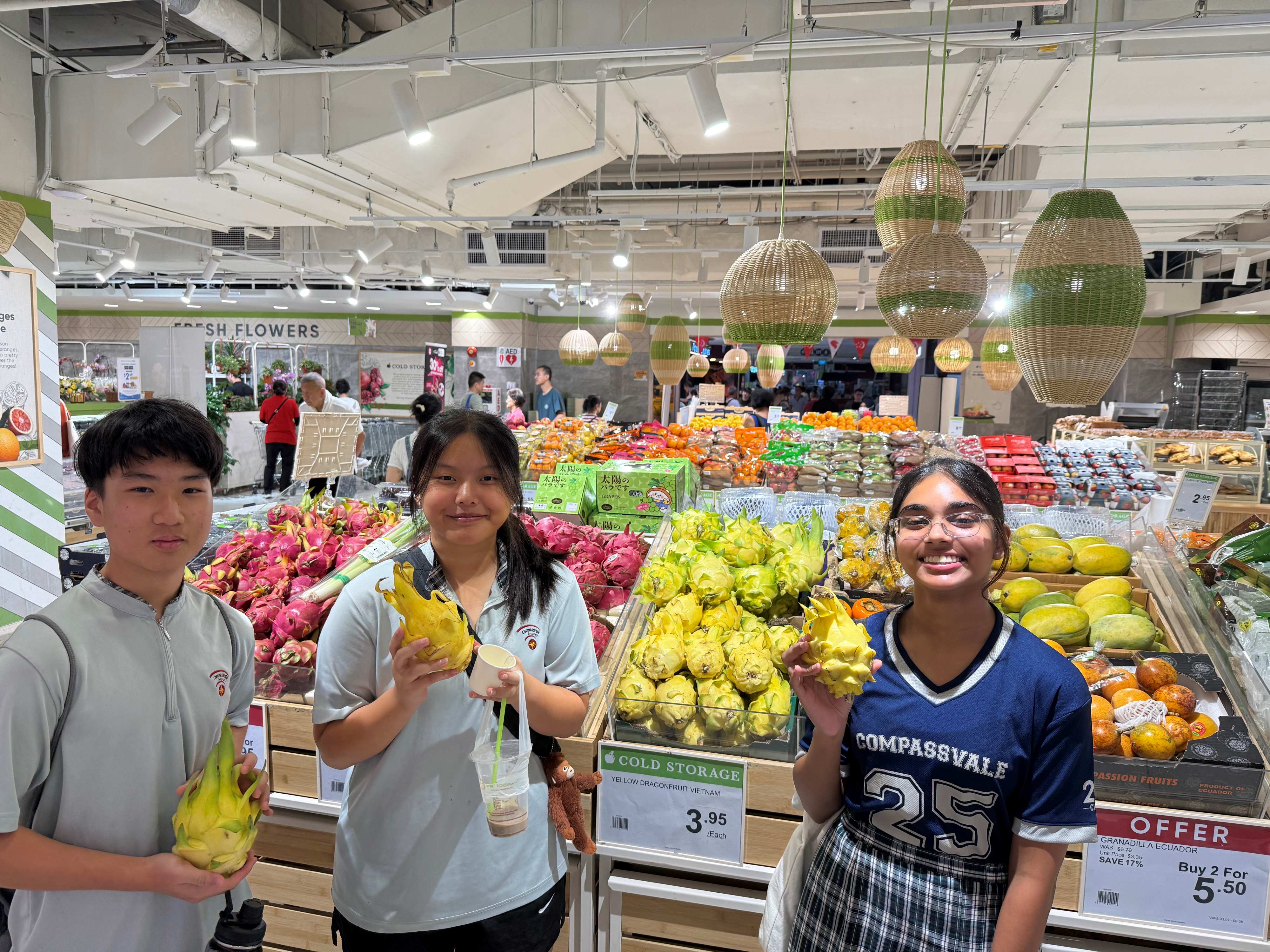 Three teenagers hold yellow dragonfruit in a Cold Storage supermarket surrounded by colorful fruit displays.