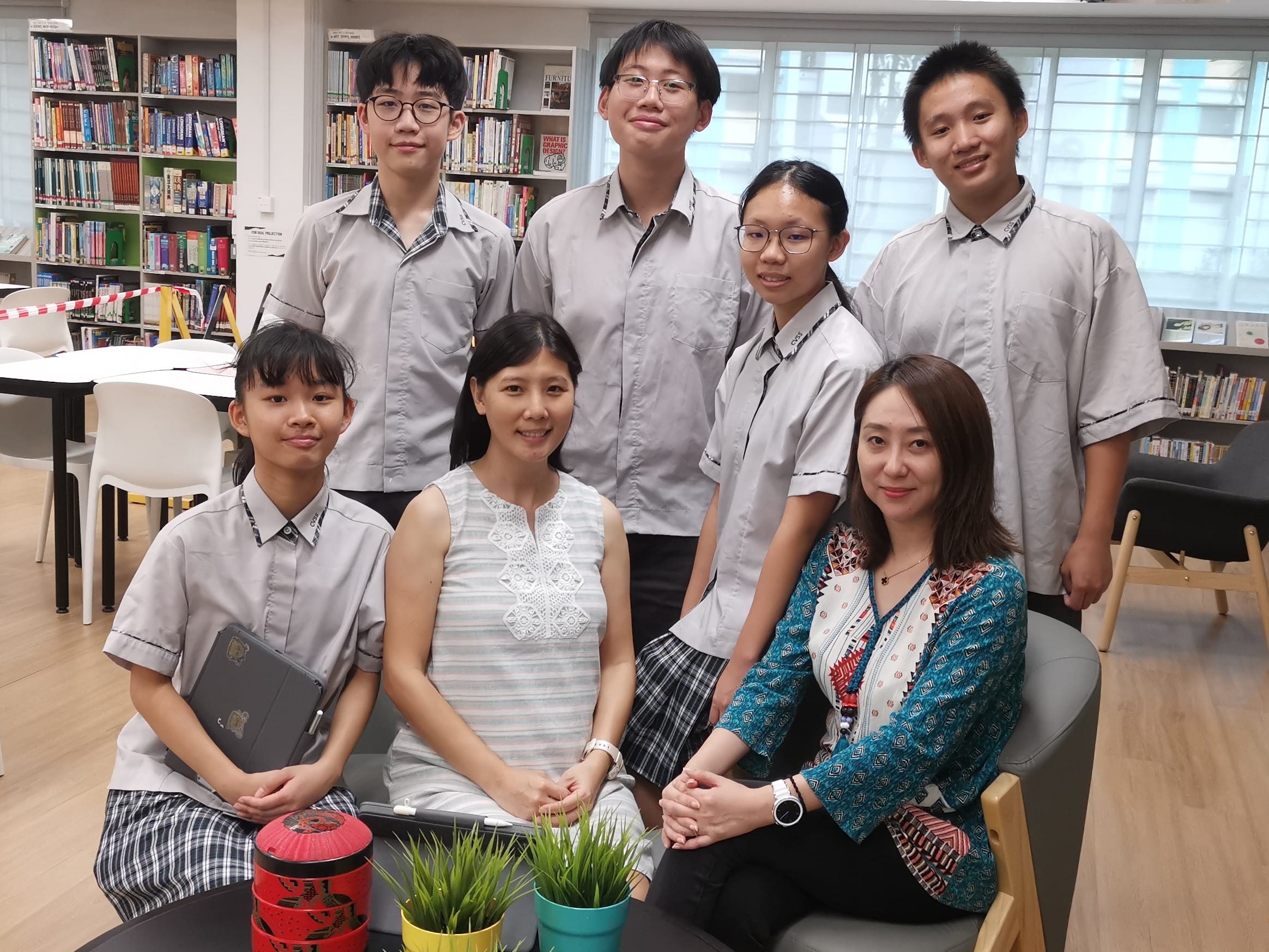 Seven people, including five students in grey uniforms and two adults, pose together smiling in a school library.