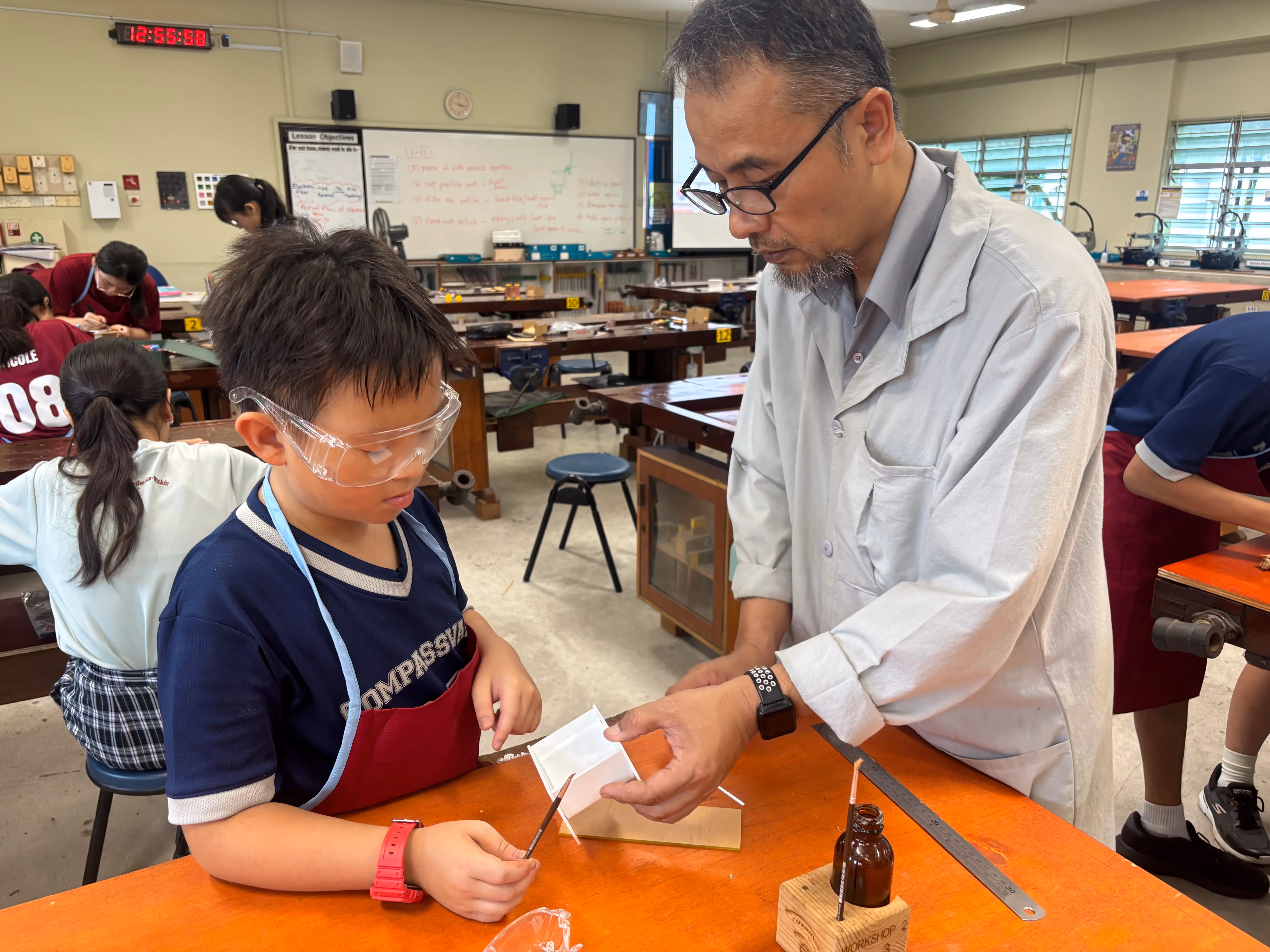 Teacher in lab coat guides student in safety goggles at a workbench in a classroom workshop.