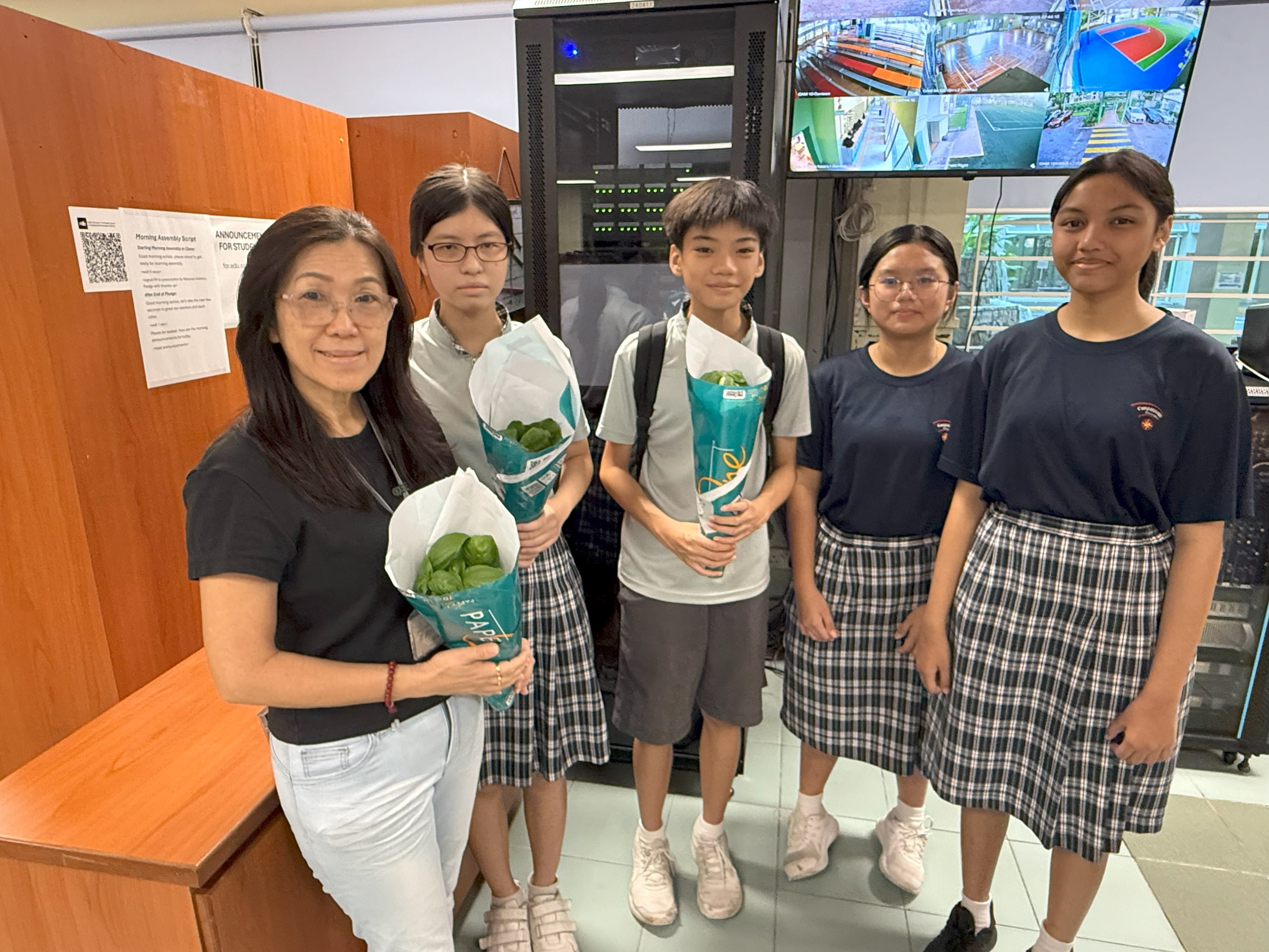 A woman and four students in school uniforms stand indoors holding wrapped green plants, with security monitors behind them.