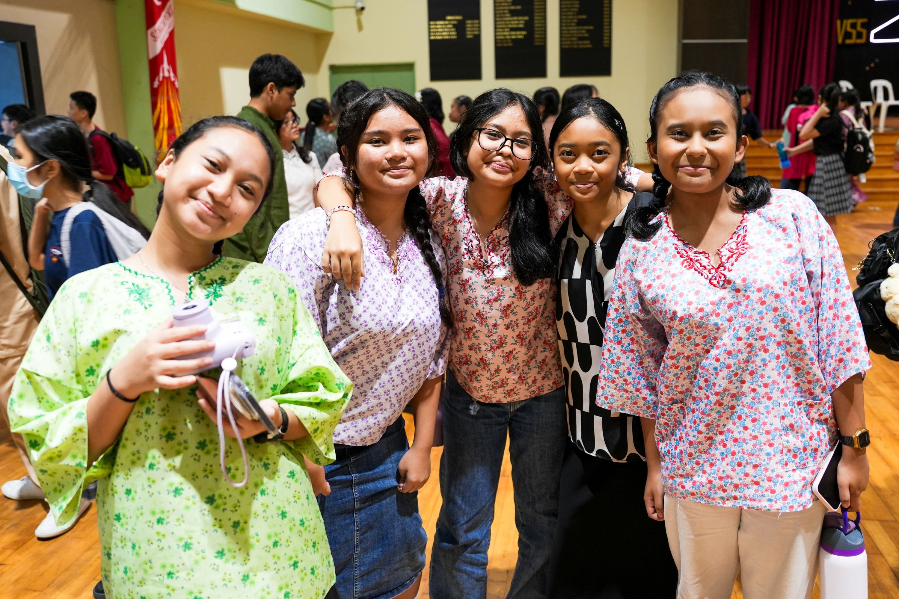 Five young girls in floral tops smile together in a school hall, one holding a small camera.