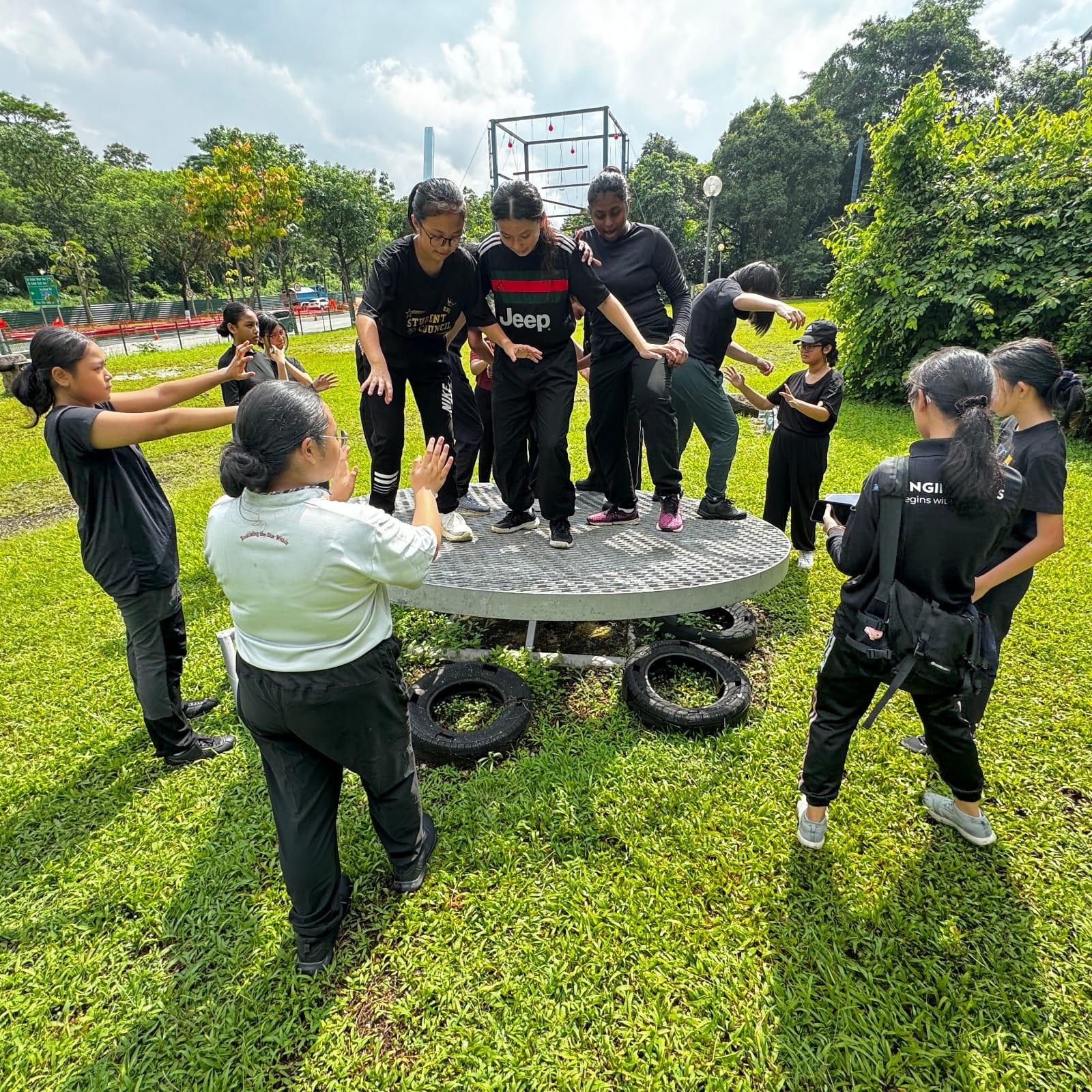 A group of young people in black clothes stand on and around a round metal platform outdoors during a team activity.