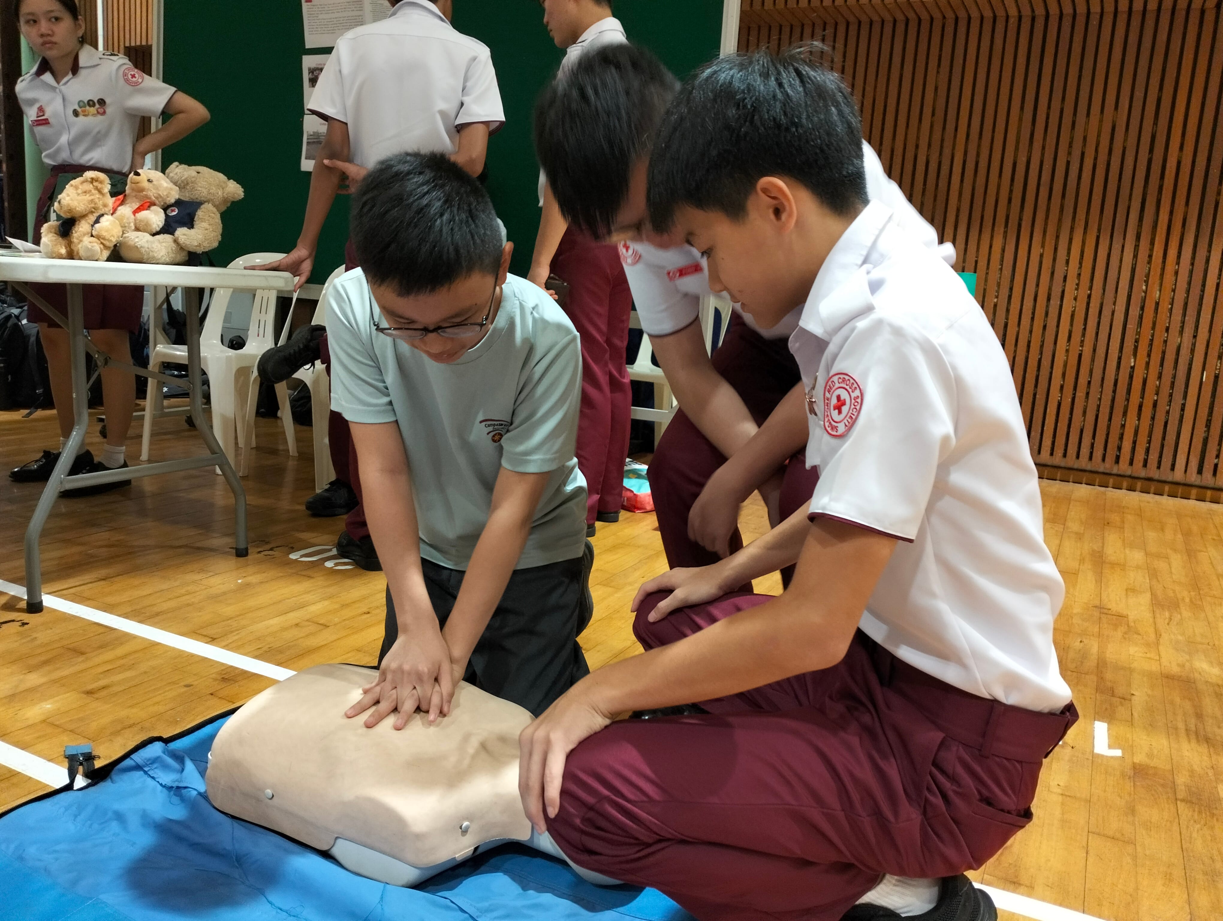 Two students practice CPR chest compressions on a training mannequin on a gym floor during a first aid session.
