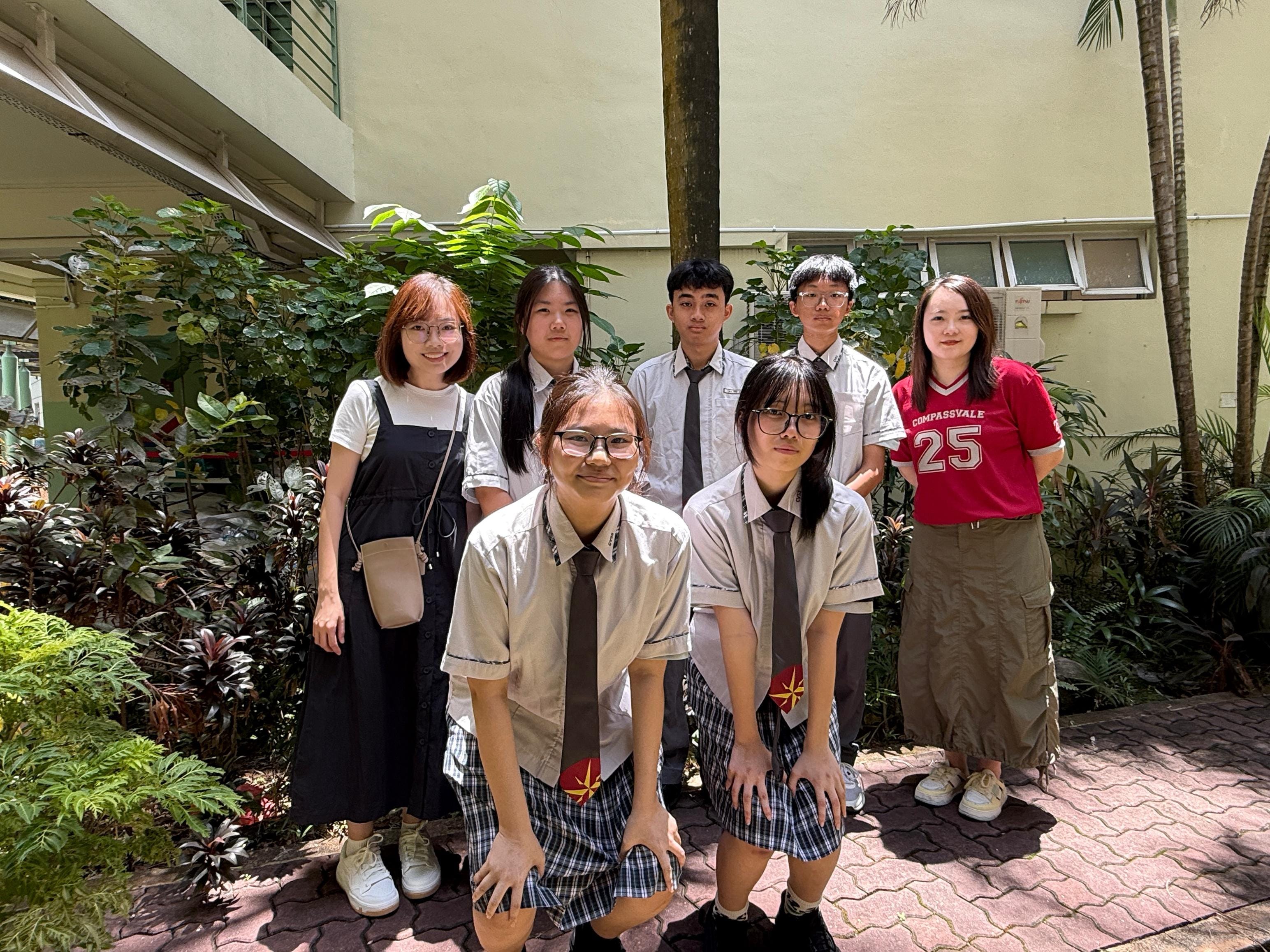 Seven young people pose outdoors near a building, some in school uniforms with ties and plaid skirts, surrounded by green plants.
