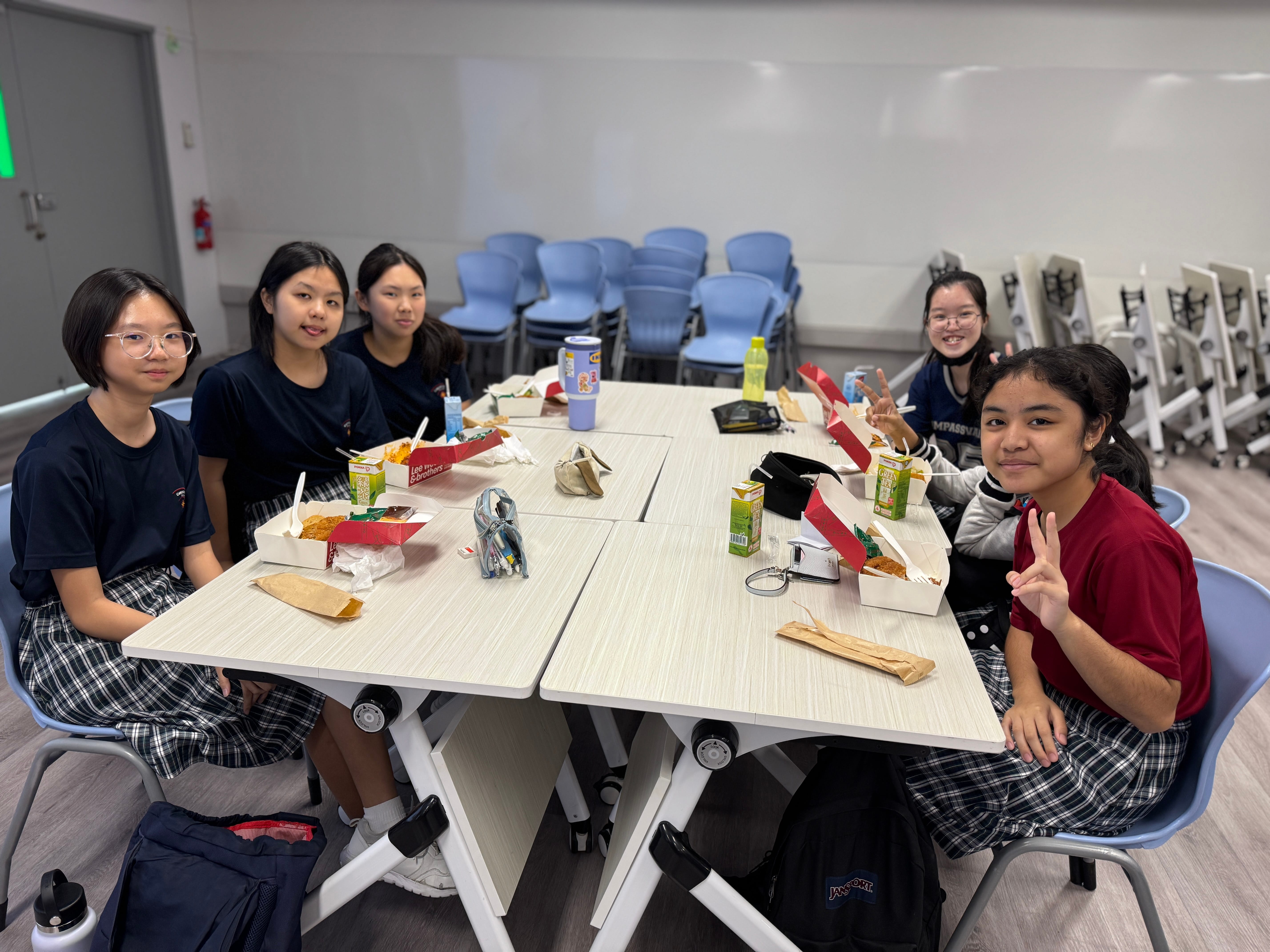 Five students sit at joined tables with takeout meals and drinks; two flash peace signs; stacked chairs behind.