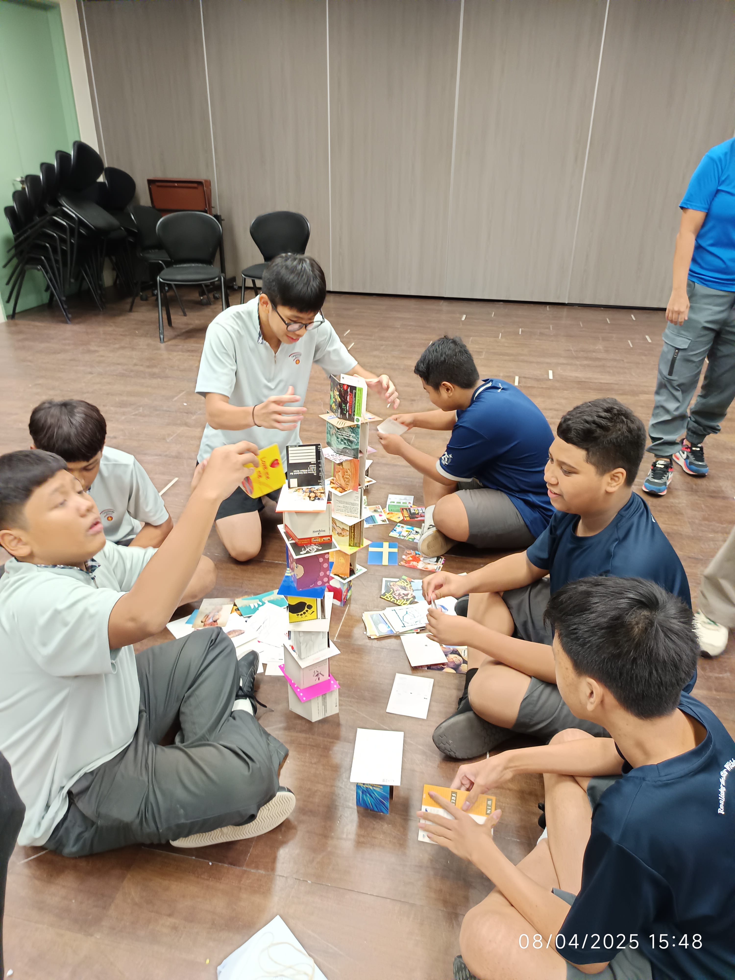 Five boys sit on a floor building a tall tower using stacked cards and small books during a group activity.