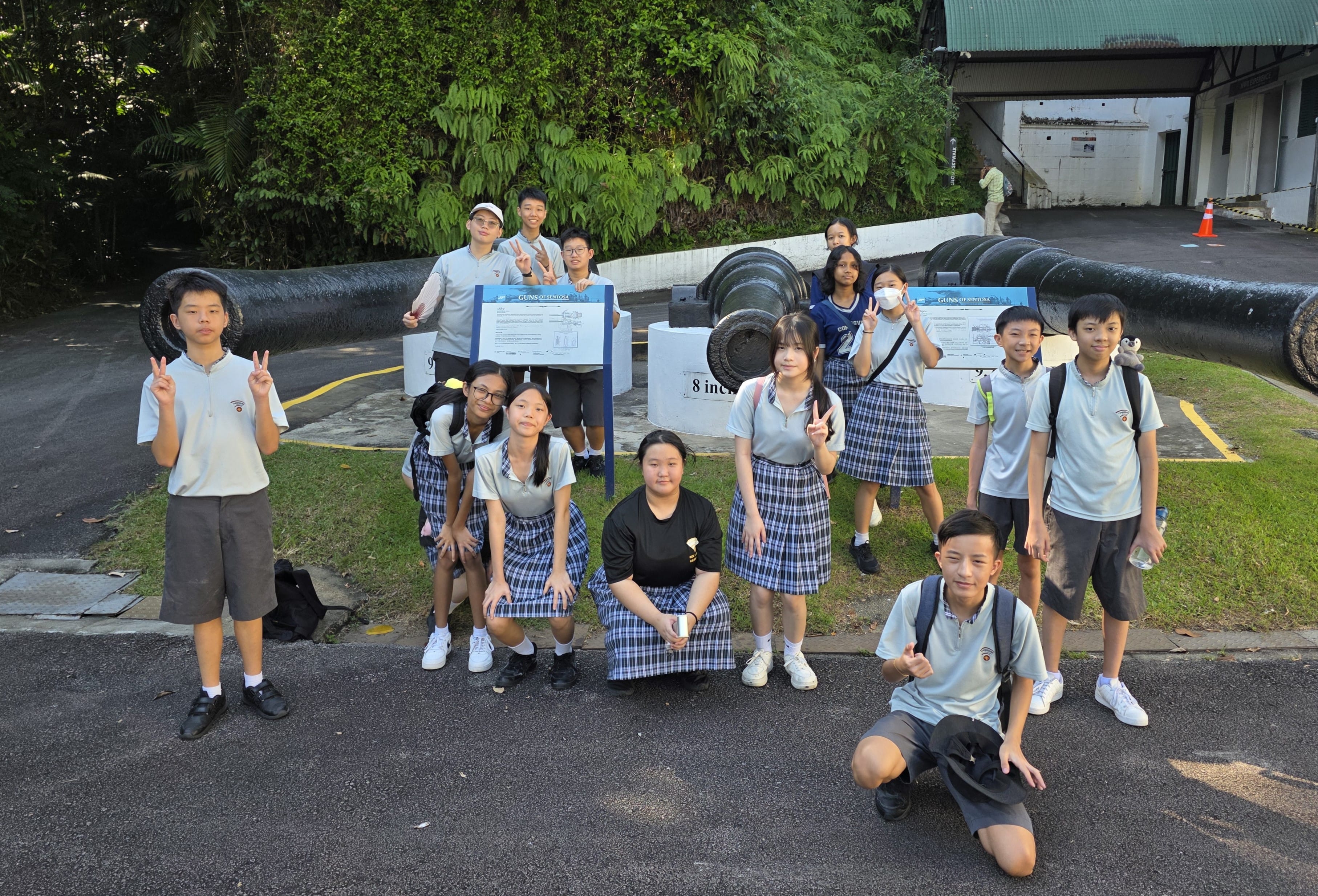 A group of uniformed students posing with peace signs in front of large historic cannons outdoors.