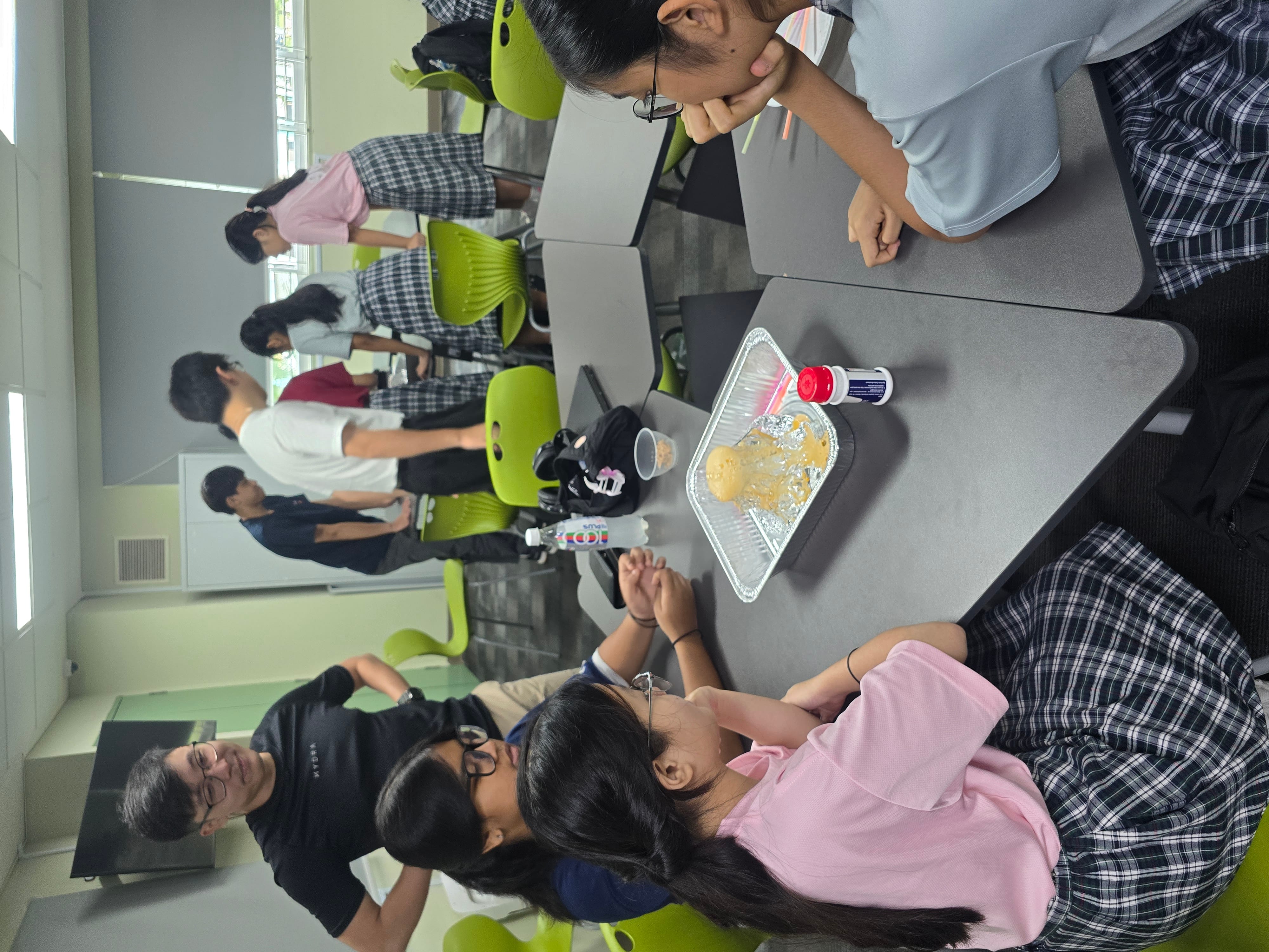 Classroom with students; two girls sit by a tray of spilled drink while others stand near the windows.