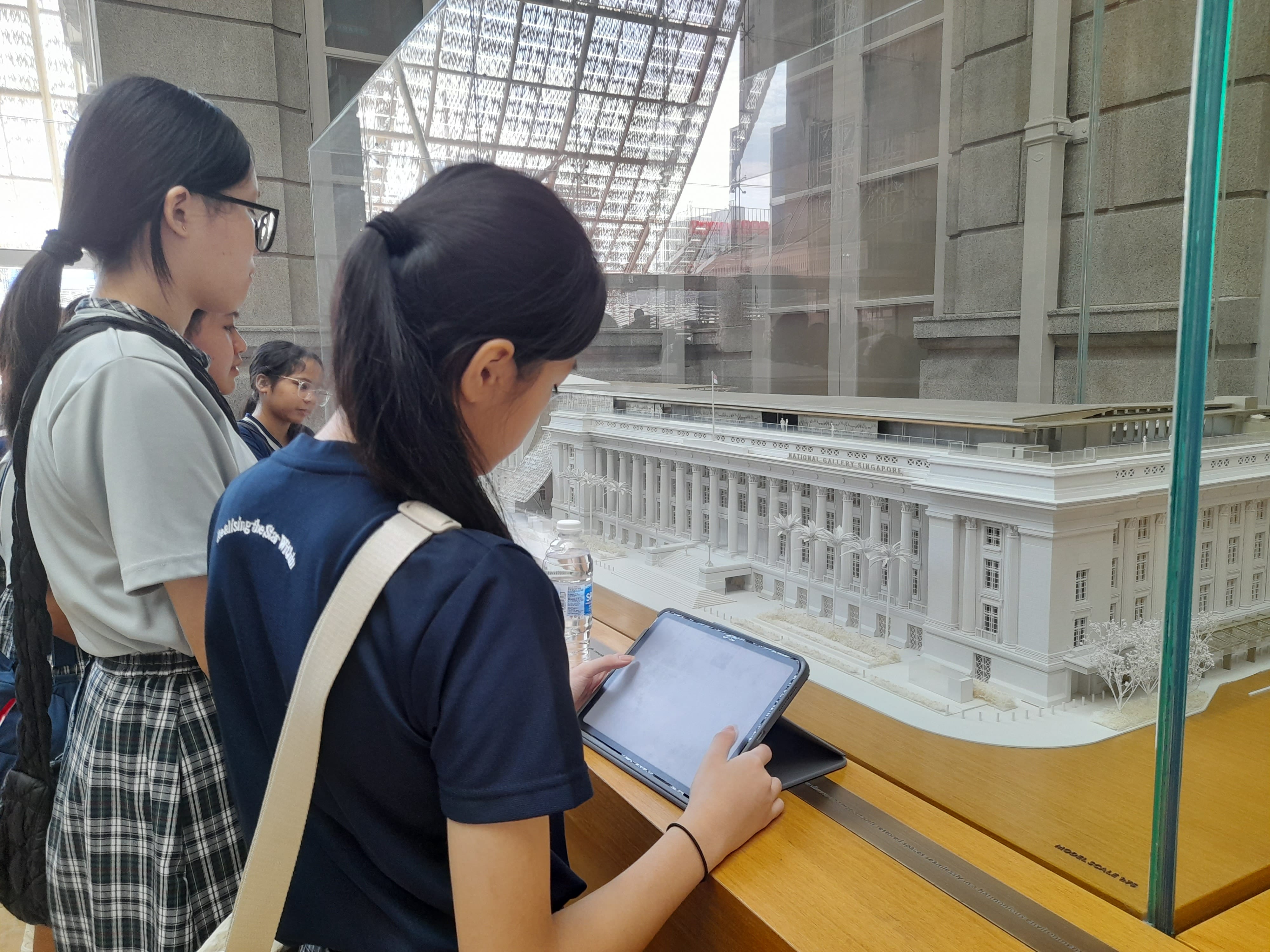 Two young women look at a detailed white architectural scale model of National Gallery Singapore, one holding a tablet.
