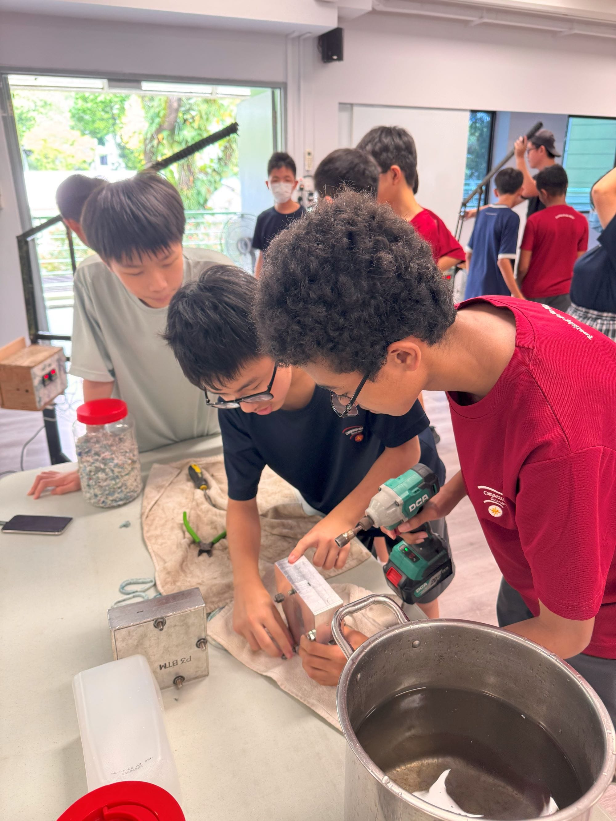 Two boys use a power drill on a workbench with tools and a metal bucket while other students work nearby.