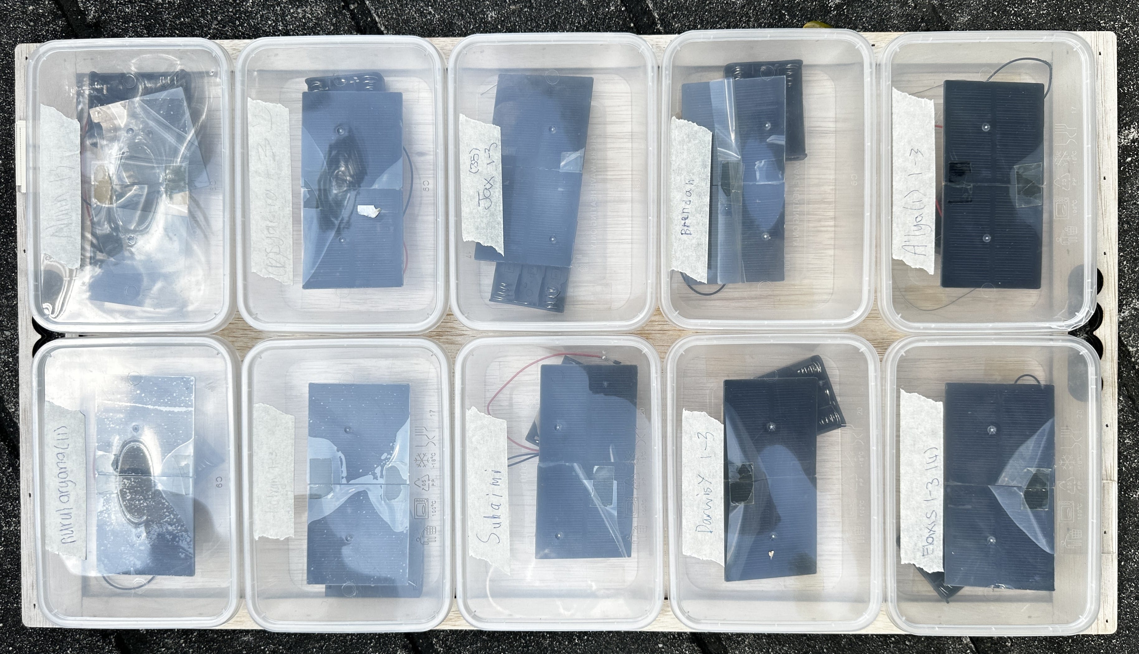 Ten clear plastic containers on a wooden board, each holding small solar panels with handwritten name labels.