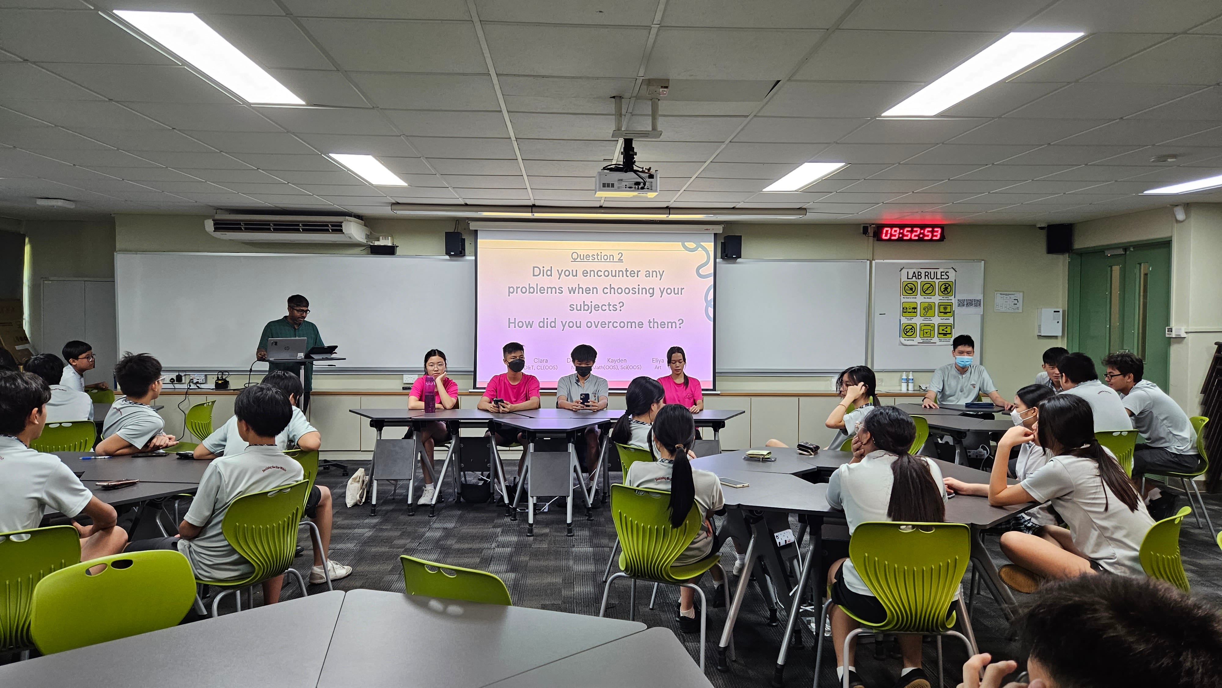 Students sit at tables facing a panel in a classroom with a projected question about choosing subjects.