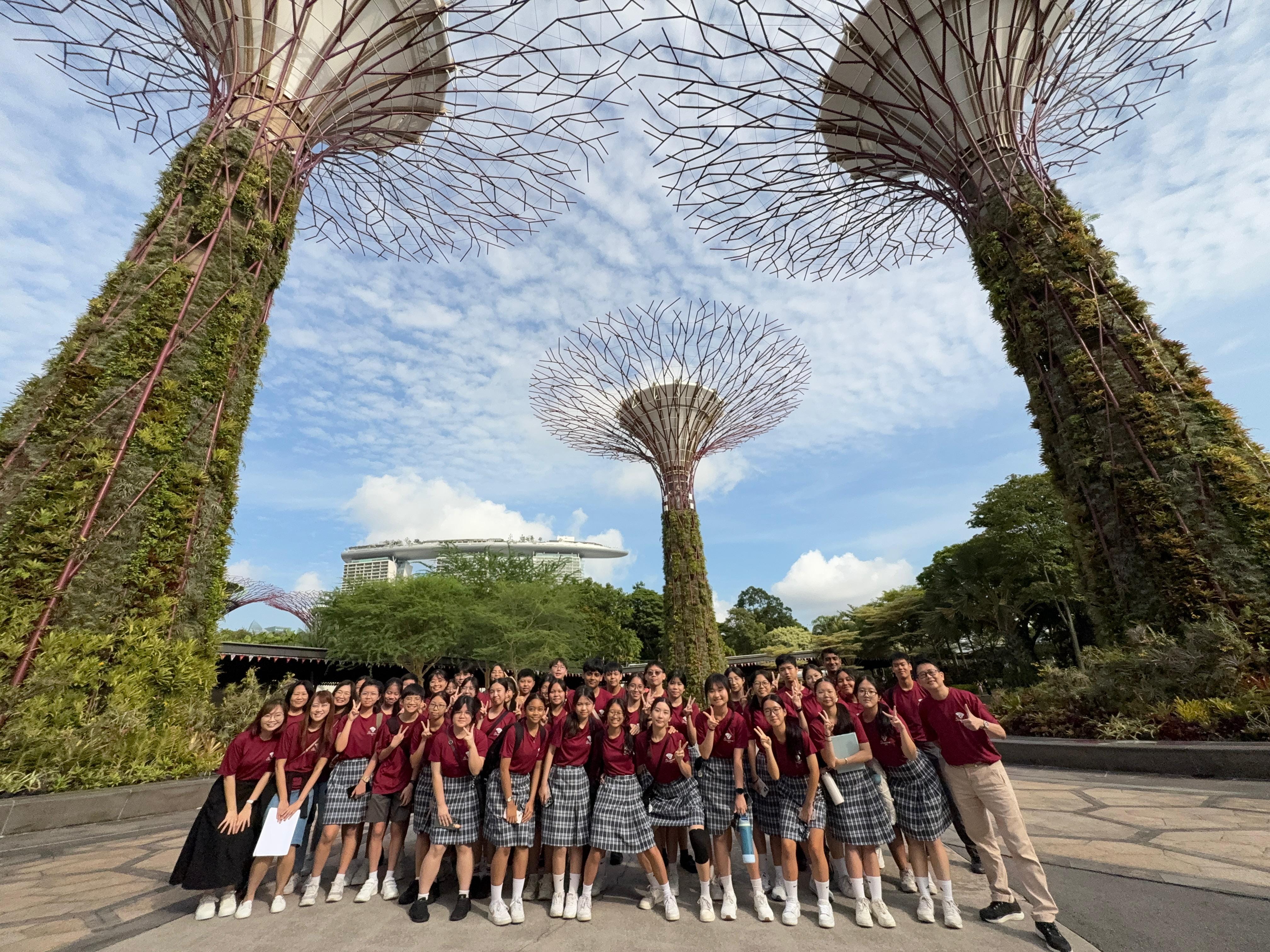 A large group of students in matching red shirts pose at Gardens by the Bay, Singapore, surrounded by Supertree structures.