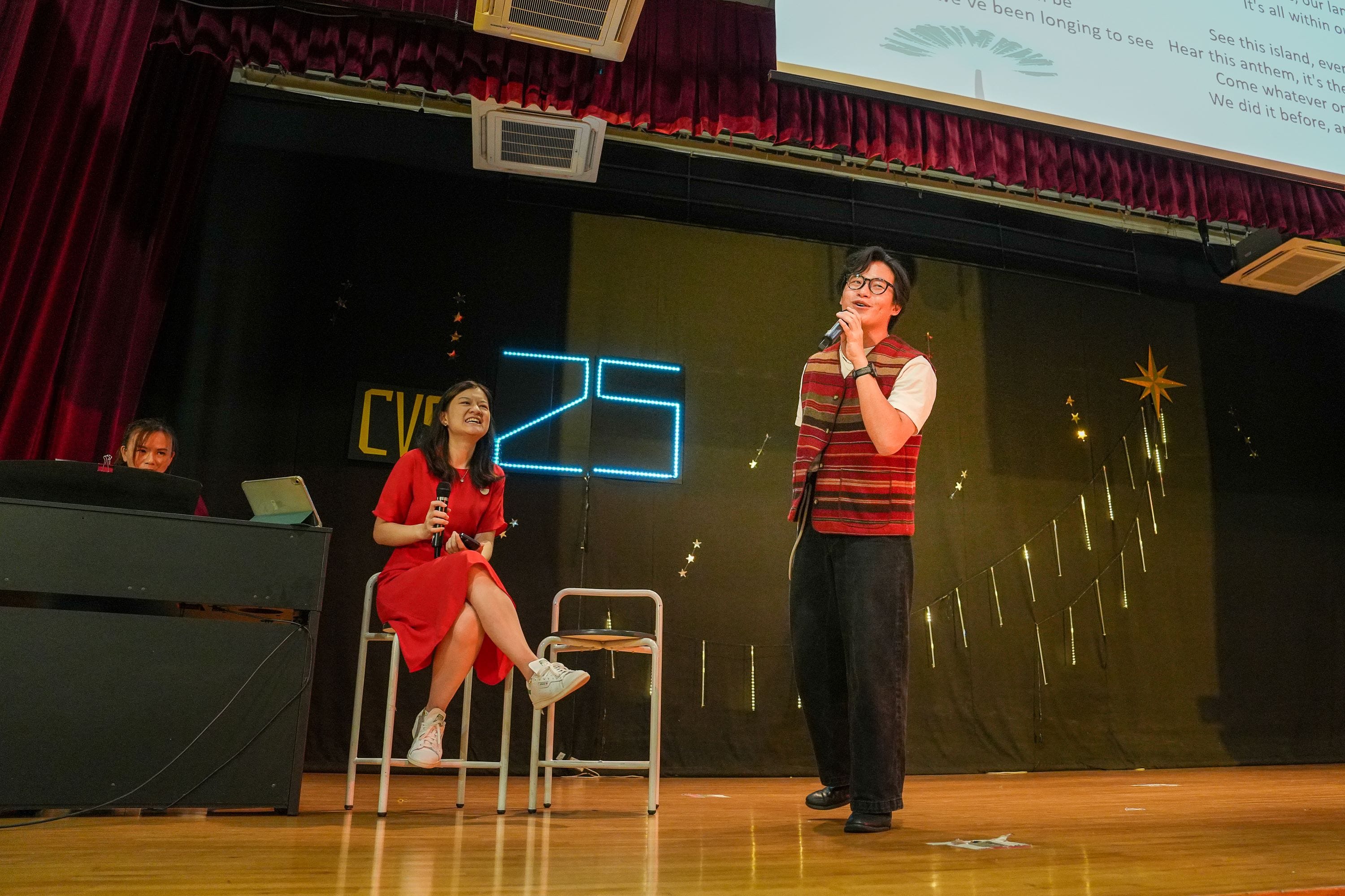 Two performers on stage, one seated in red dress holding mic, one standing singing, with a lit "25" sign behind them.